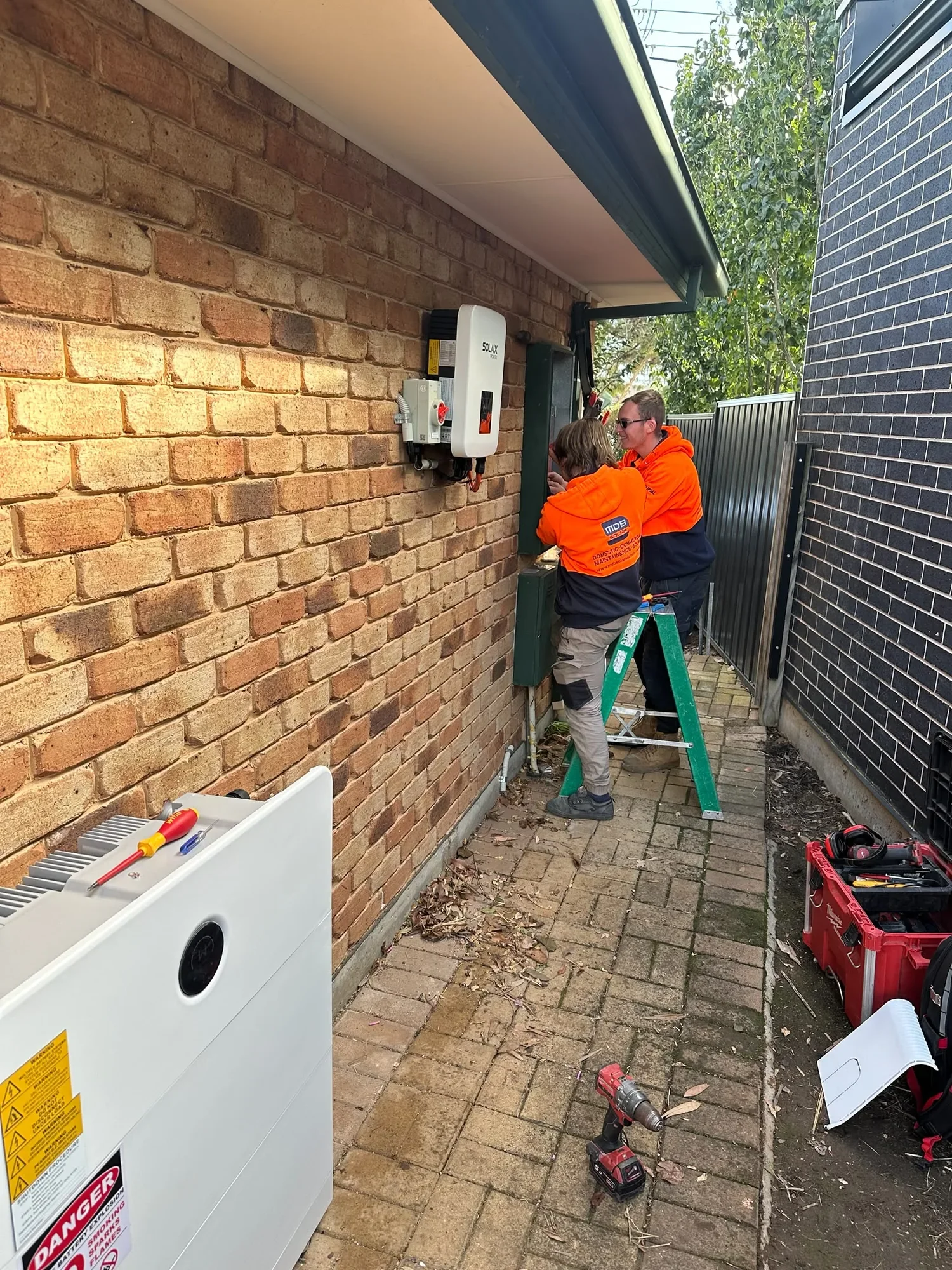 Two technicians in Orange and navy work jackets are installing an outdoor solar power inverter on a brick wall between two houses. The area has tools and a power drill on the ground, with a partly disassembled outdoor heater or boiler nearby.