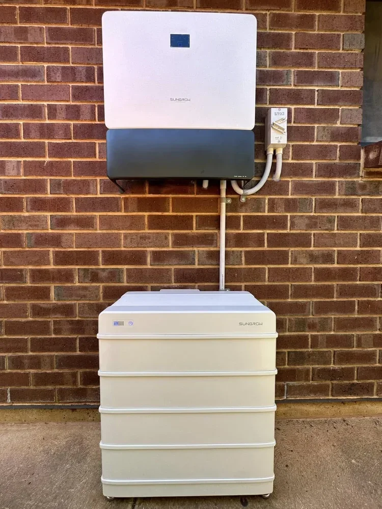 A solar power inverter system installed on a brick wall with white and gray components, connected to a white multi-drawer battery storage unit below.