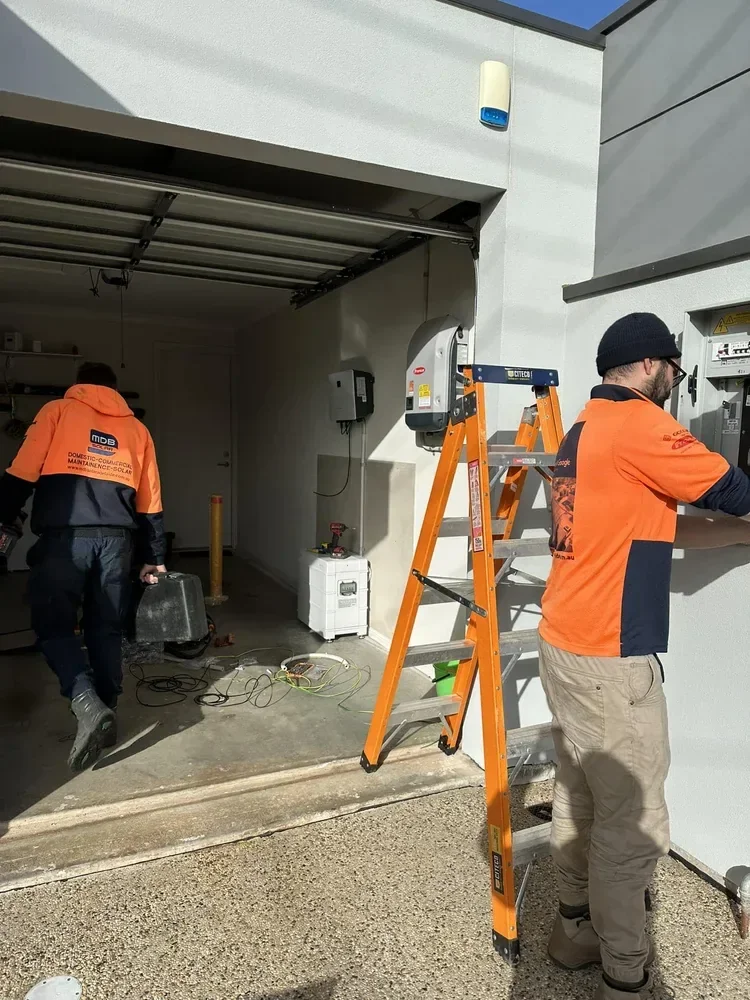 Two workers in orange and navy uniforms working outside a building with an open garage door. One worker, wearing a black beanie, is using an electrical panel, and the other, walking into the garage, carries equipment. An orange ladder is propped agai