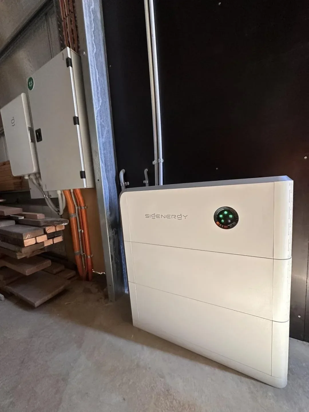 A white solar energy system unit with a circular control panel on the front, labeled 'SIGENERGY', placed on a concrete floor near electrical wiring and an electrical box.