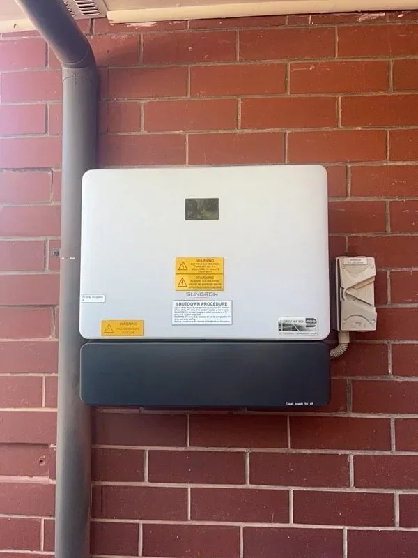 An outdoor solar inverter mounted on a brick wall with warning labels, a gray conduit pipe on the left, and a small gray electrical box on the right.