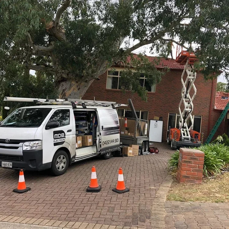A white service van parked on a brick driveway with orange traffic cones in front. The van has its side door open, revealing tools and supplies inside. Behind the van, there is a tall tree with a branch extending over the house. A cherry picker lift 