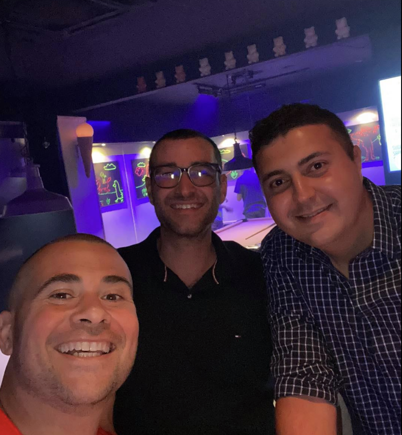 Three men smiling and taking a selfie in a dimly lit bar or restaurant with neon artwork and television screens in the background.