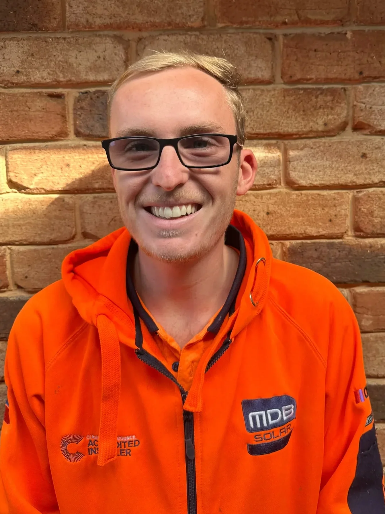 A young man with blond hair, glasses, and a goatee smiling, wearing an orange jacket with black accents, standing against a brick wall.