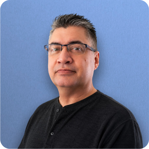 A man with short dark hair, glasses, and a serious expression, wearing a black shirt, stands against a blue wall.