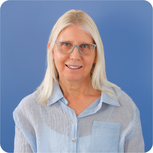 A smiling elderly woman with blonde hair and glasses, wearing a light blue button-up shirt against a blue background.