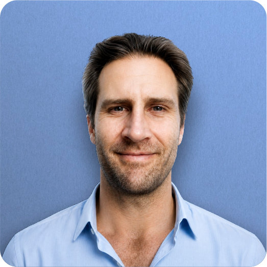 A man with dark brown hair and a beard, wearing a light blue collared shirt, smiling in front of a blue background.