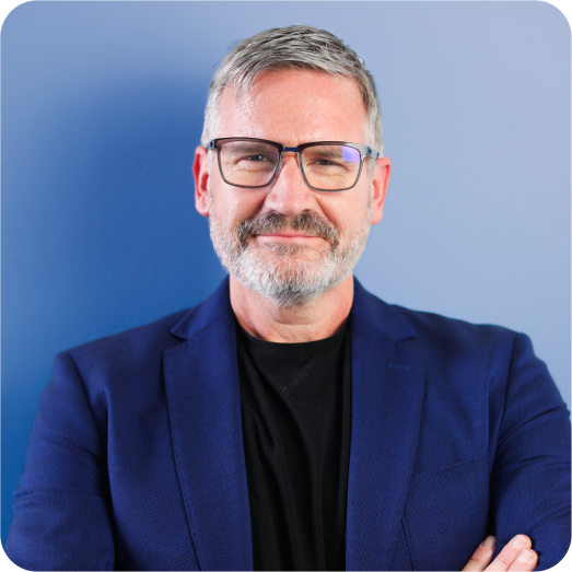 A middle-aged man with gray hair and a beard, wearing glasses, a black shirt, and a navy blue blazer, standing against a blue background and smiling.