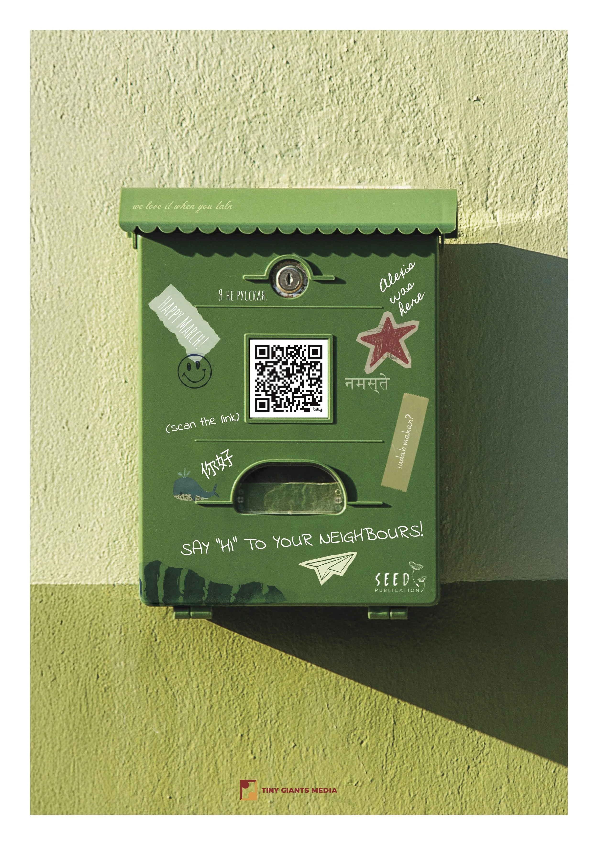 Green mailbox covered in stickers and handwritten notes, mounted on a wall with a shadow cast to its right. The stickers include a happy face, a star, a whale, and various messages in different languages. The wall is textured and beige. There is a QR