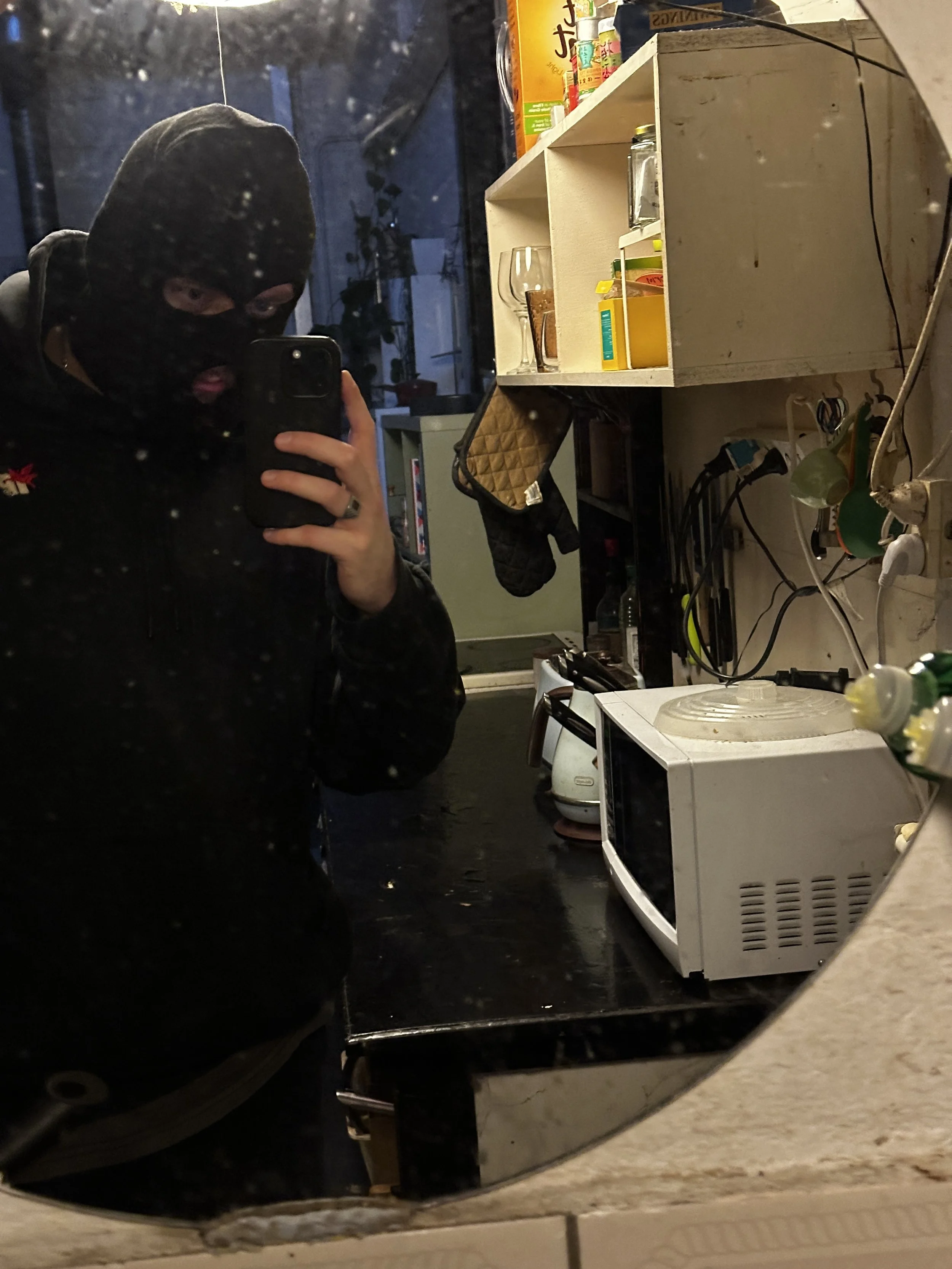A person wearing a black ski mask taking a selfie in a mirror in a cluttered room with kitchen appliances, a computer, and various items on shelves.