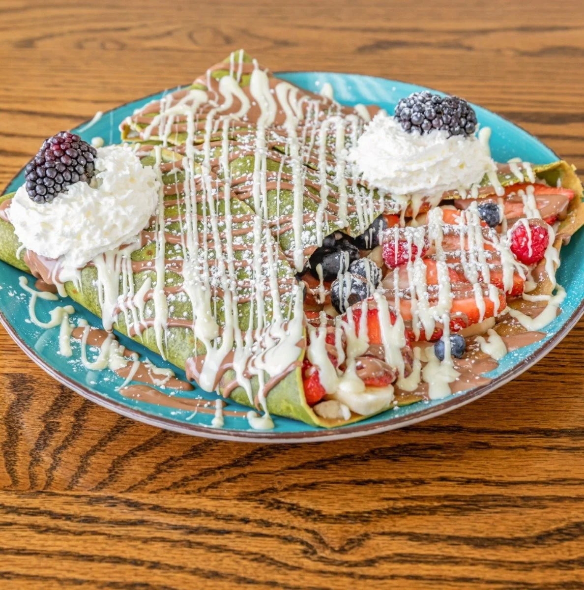 Colorful dessert crepe topped with white and milk chocolate drizzle, whipped cream and blackberries, with additional strawberries, blueberries, and raspberries inside, served on a blue plate on a wooden table.