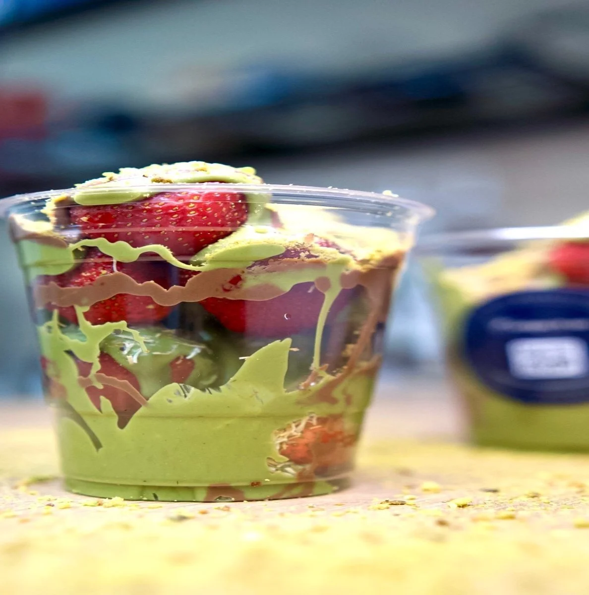 Clear plastic cup filled with fresh strawberries and green lettuce, topped with dressing, placed on a yellow textured surface, with a blurred background of a condiment container.