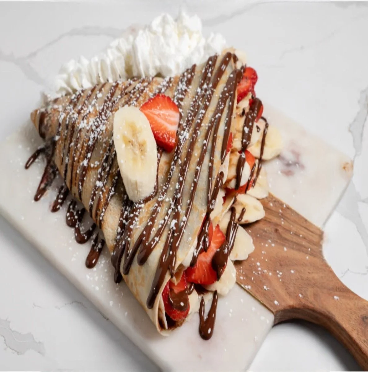 A crepe filled with sliced bananas, strawberries, and whipped cream, drizzled with chocolate sauce, dusted with powdered sugar, served on a wooden paddle-shaped plate.