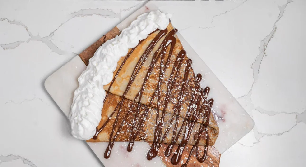 A dessert crepe topped with chocolate sauce, powdered sugar, and whipped cream, served on a white marble surface.
