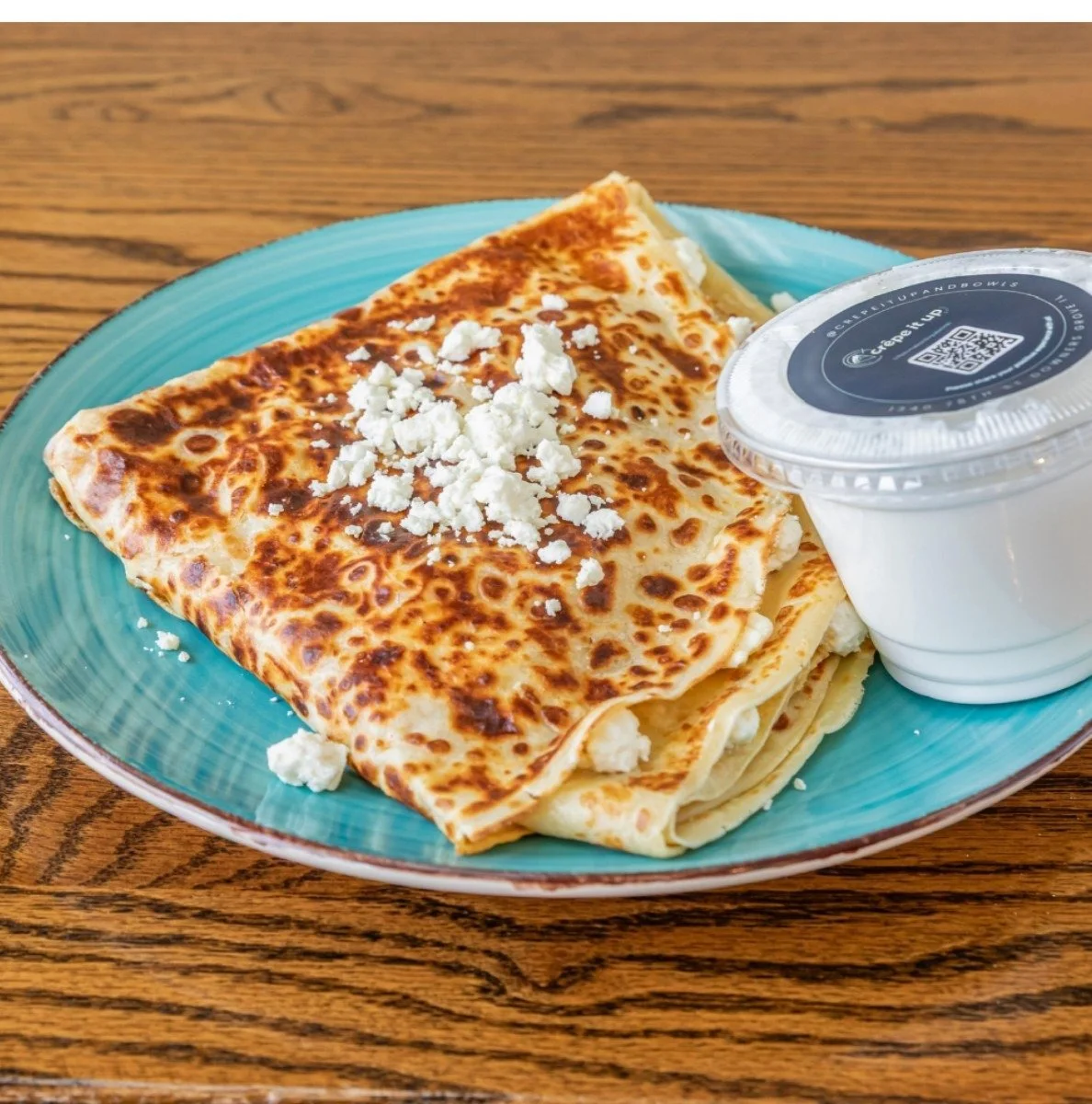 Cheese quesadilla with crumbled cheese on top, served on a green plate with a side of sour cream in a plastic cup, on a wooden table.