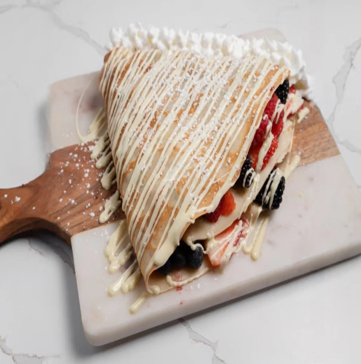 A slice of berry-filled crepe drizzled with white chocolate on a wooden board.