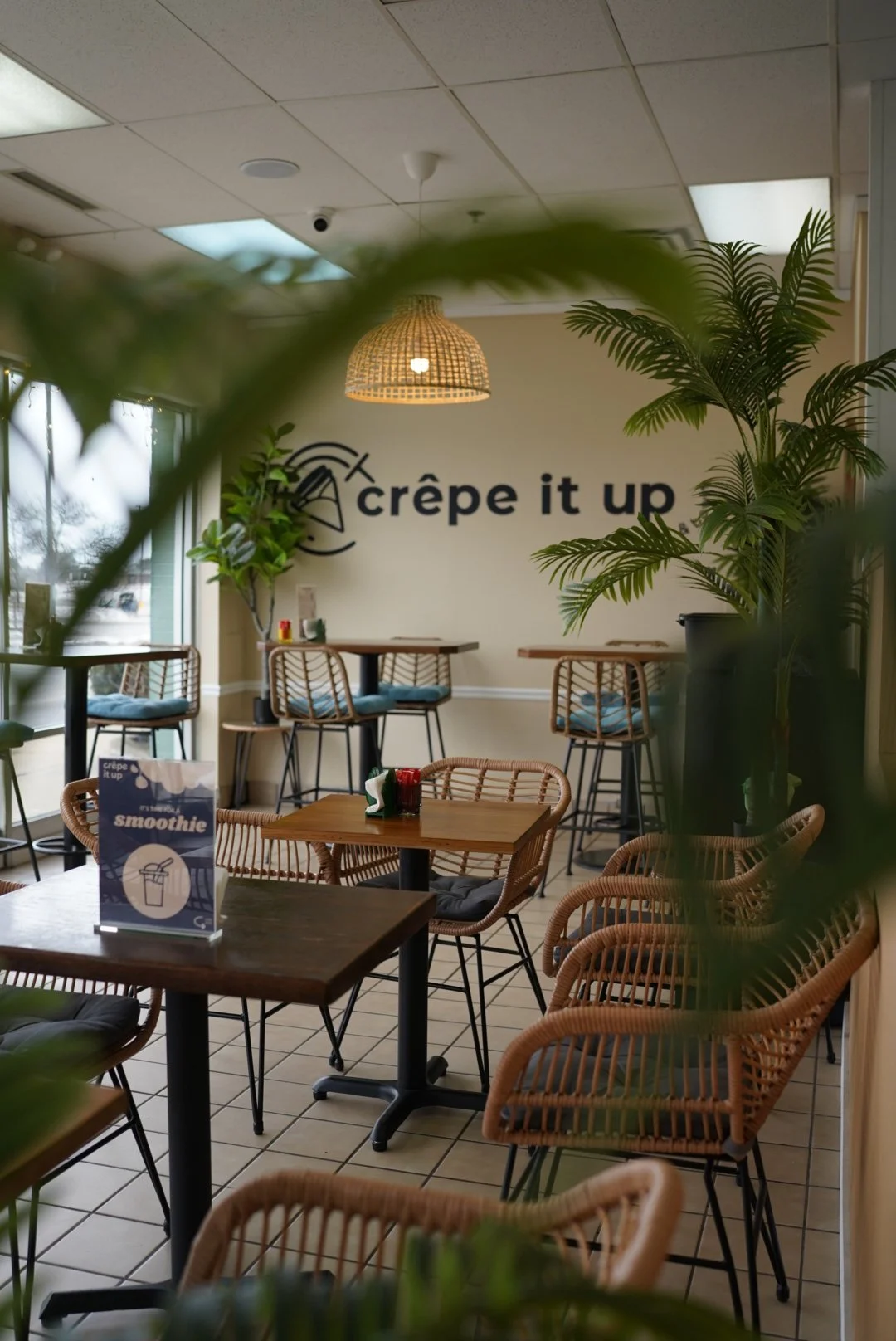 Interior of a cozy cafe with wicker chairs, wooden tables, a sign reading 'crêpe it up,' and large windows with greenery outside.