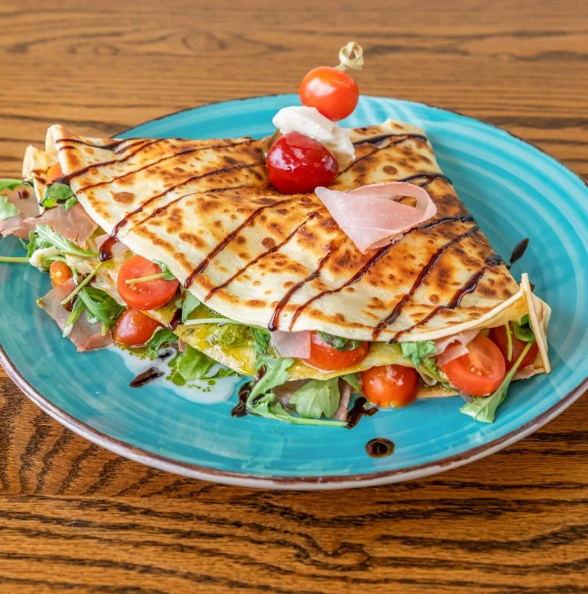 A plate with a folded crepe filled with vegetables, cherry tomatoes, and ham, garnished with cherry tomatoes and whipped cream on top, drizzled with balsamic glaze.