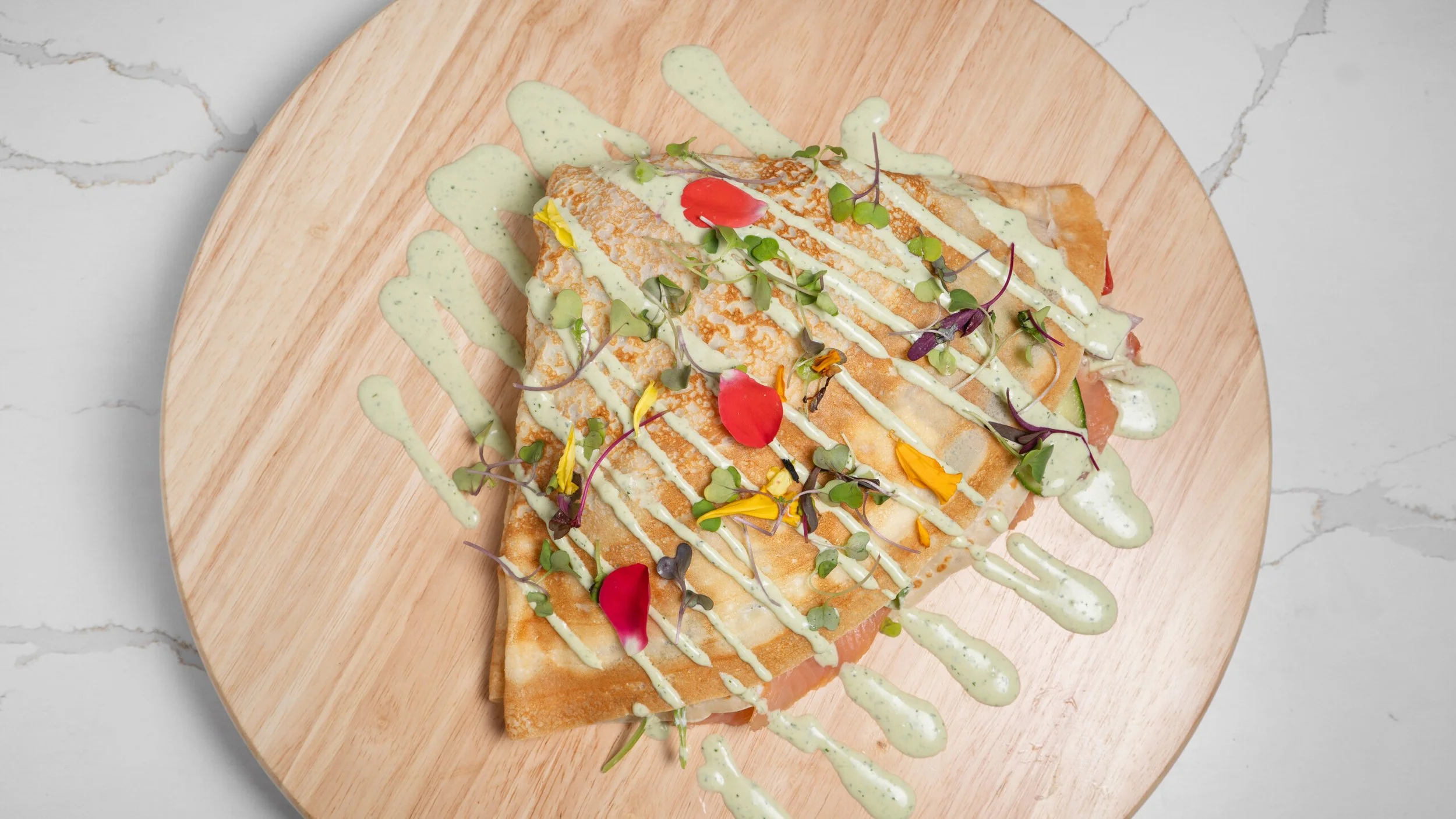 A savory crepe topped with microgreens, edible flowers, and drizzled with green herb sauce and a white creamy sauce, served on a wooden plate.