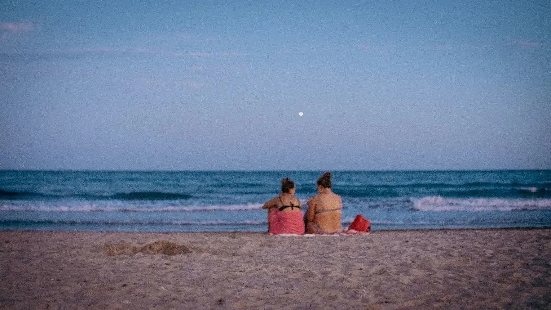 Two people sitting on the beach looking toward the horizon in Between Two Shores.