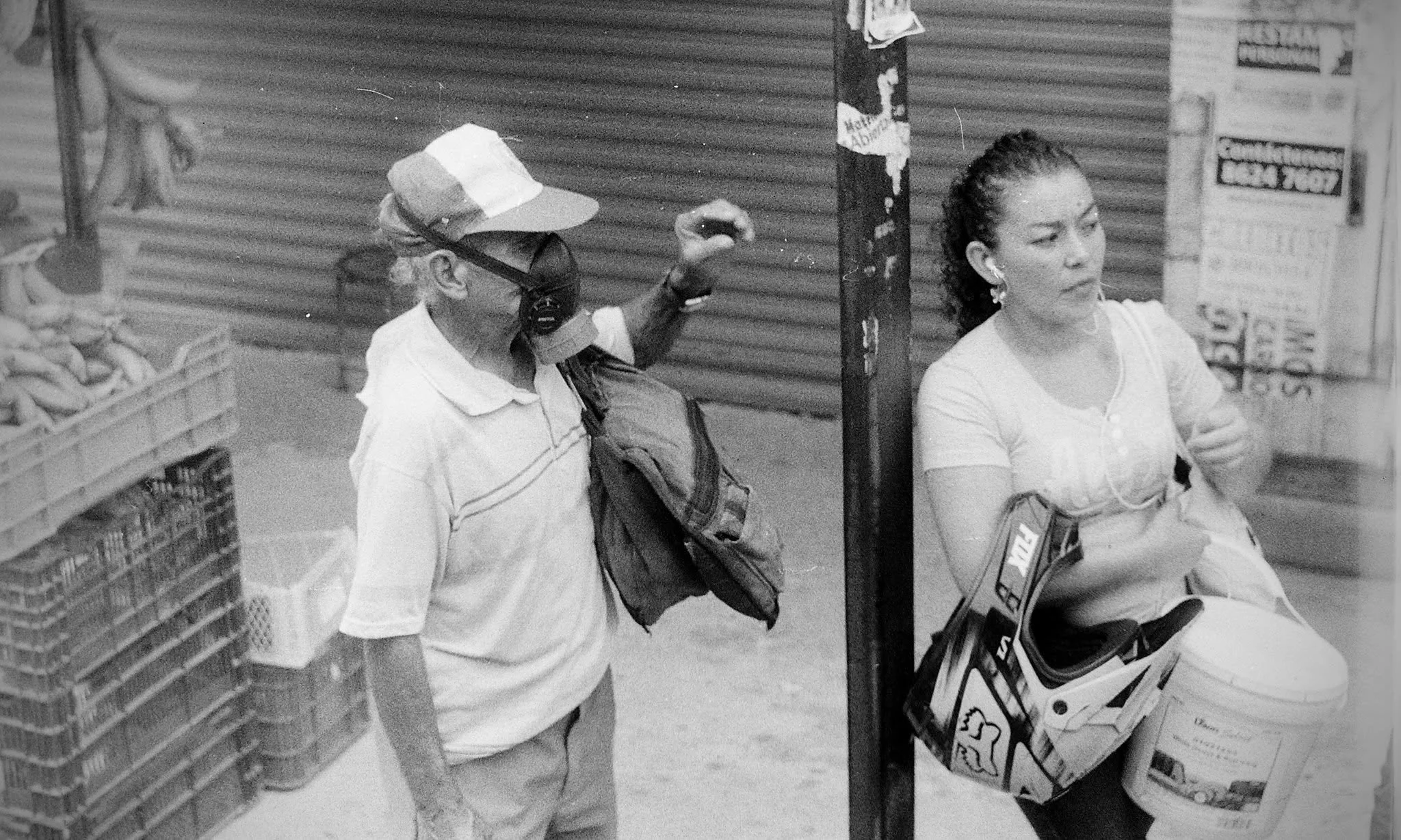 A woman with curly hair in a white shirt sitting next to a man with glasses, a hat, and a face mask, who is holding a phone and raising his hand. They are at a street market with closed shop shutters and baskets of produce around.