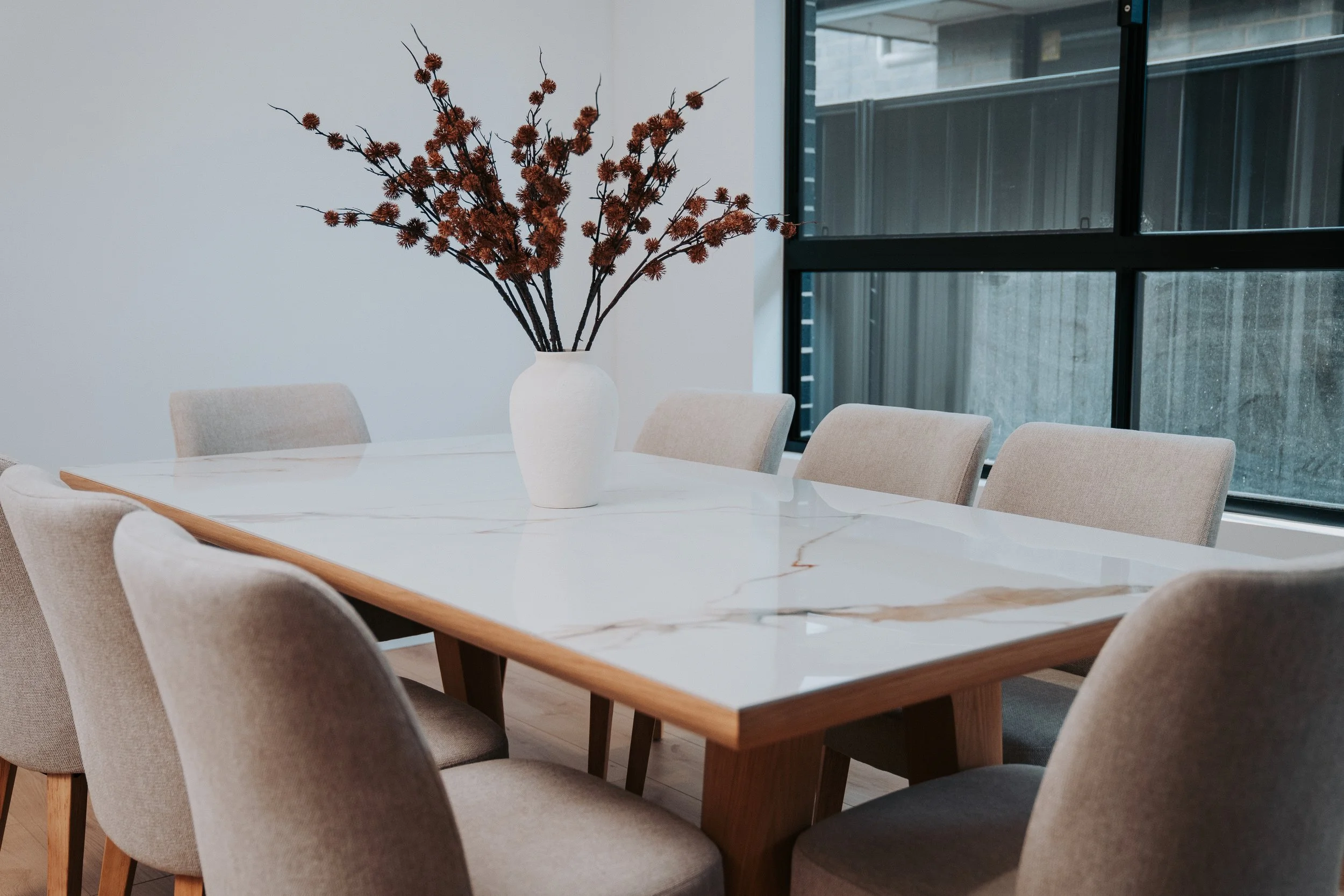 A white vase with brown artificial flowers on a white marble dining table, surrounded by beige chairs in a modern dining room with large windows.