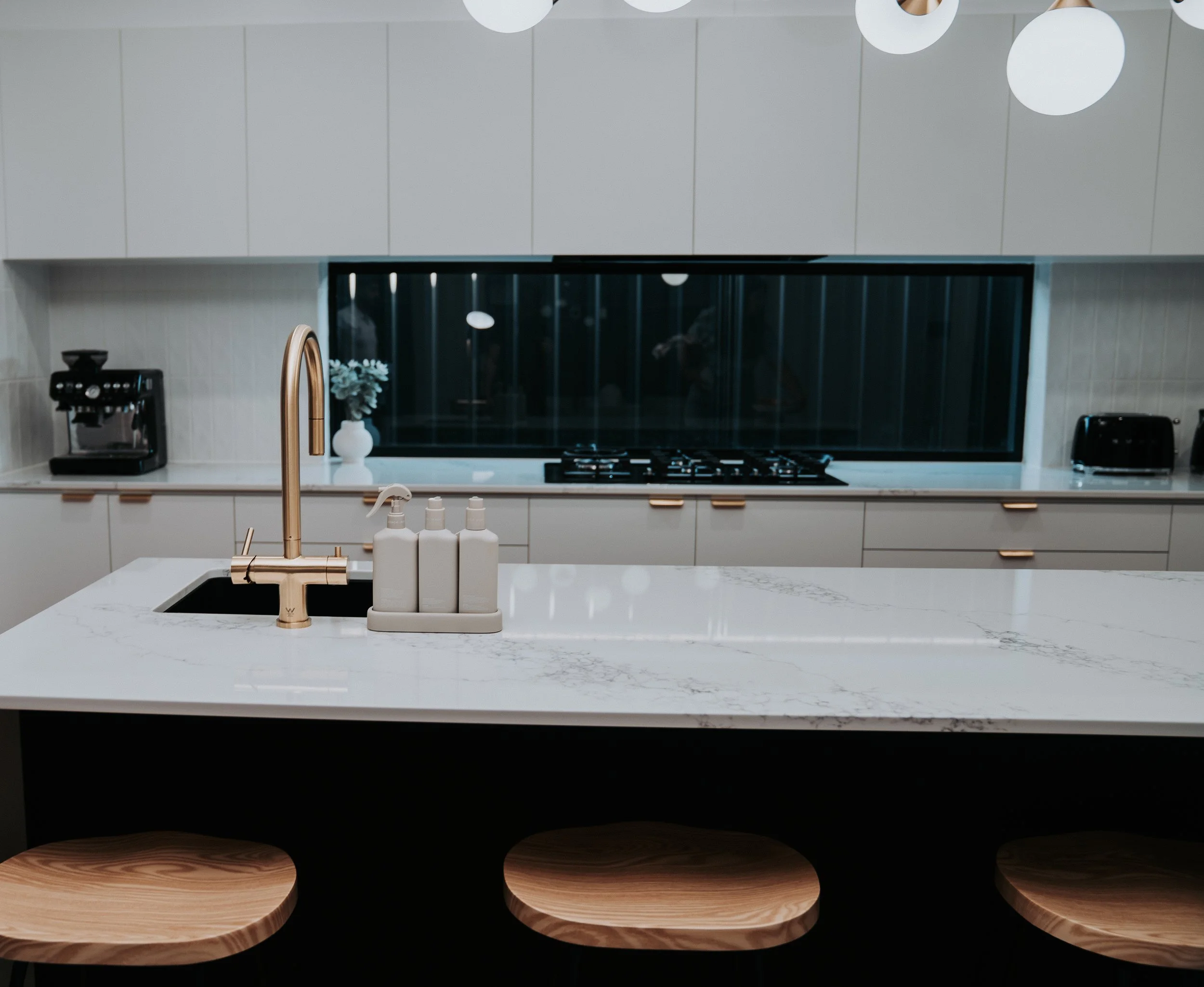 Modern kitchen with white marble island, gold faucet, black sink, beige cabinetry, and bar stools.