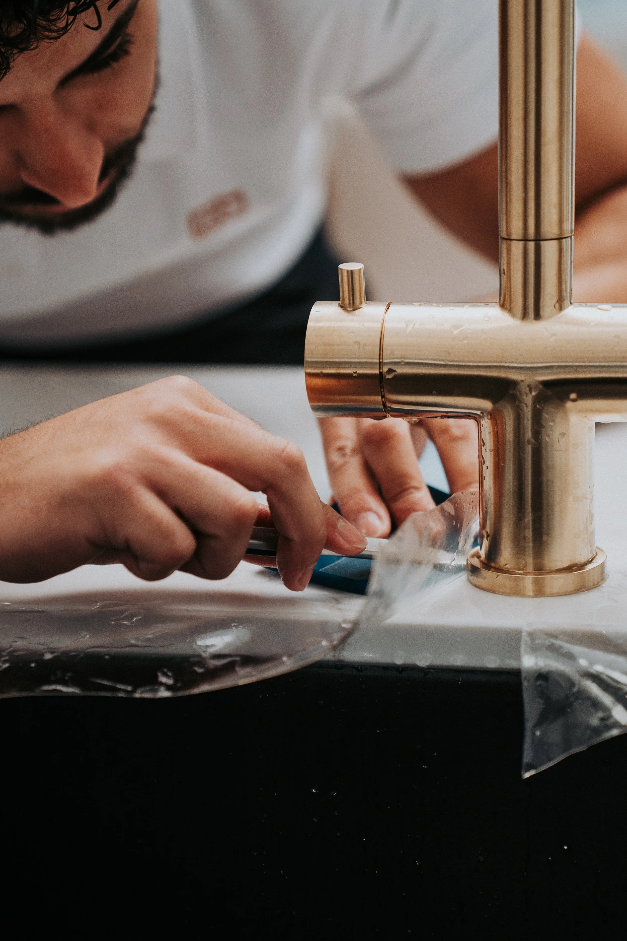 A person working on a project with a metallic copper-colored faucet being installed or repaired, with hands manipulating a tool or material on a white surface covered in plastic.