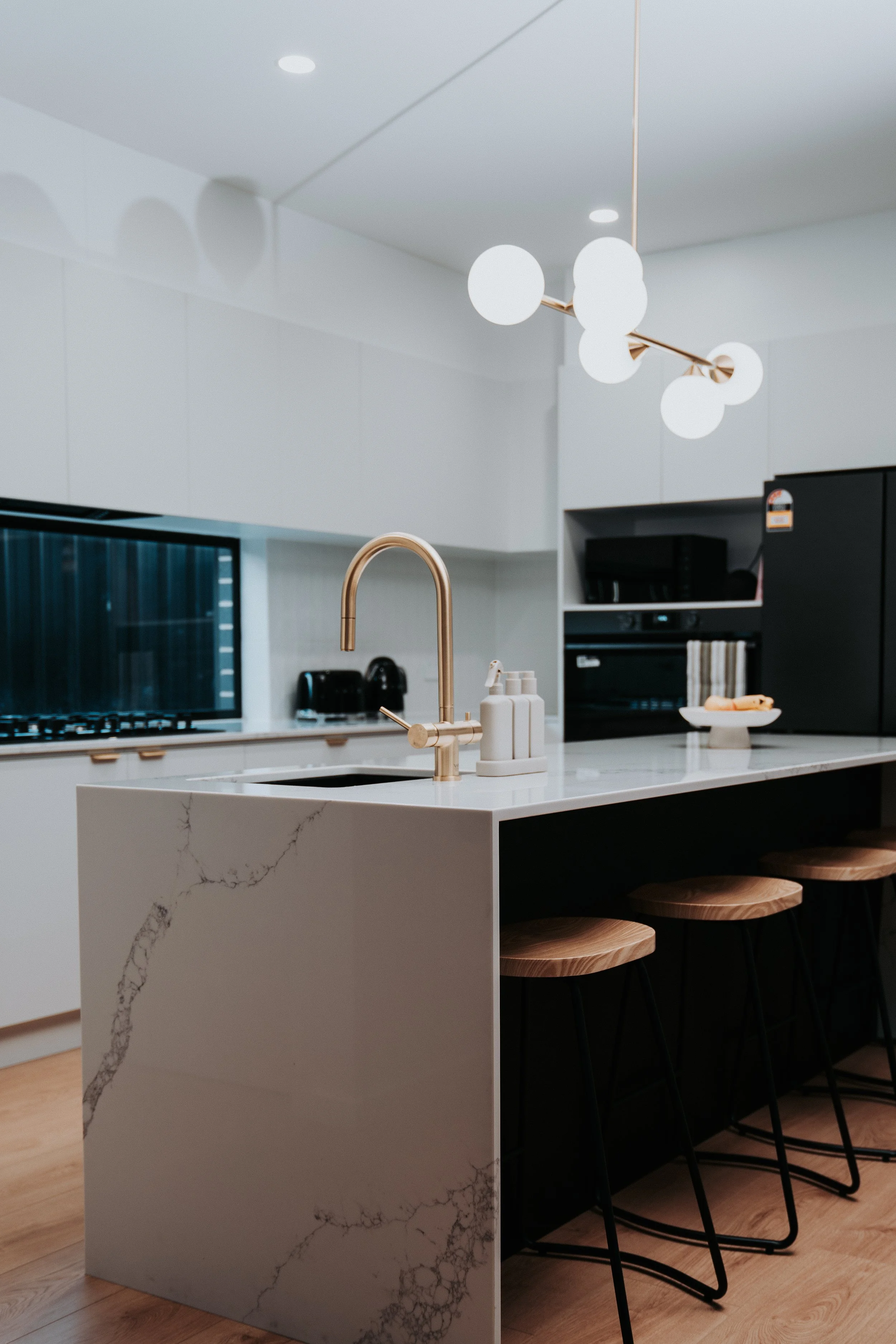 Modern kitchen with marble waterfall island, copper pendant lights, black bar stools, white cabinetry, and wooden flooring.