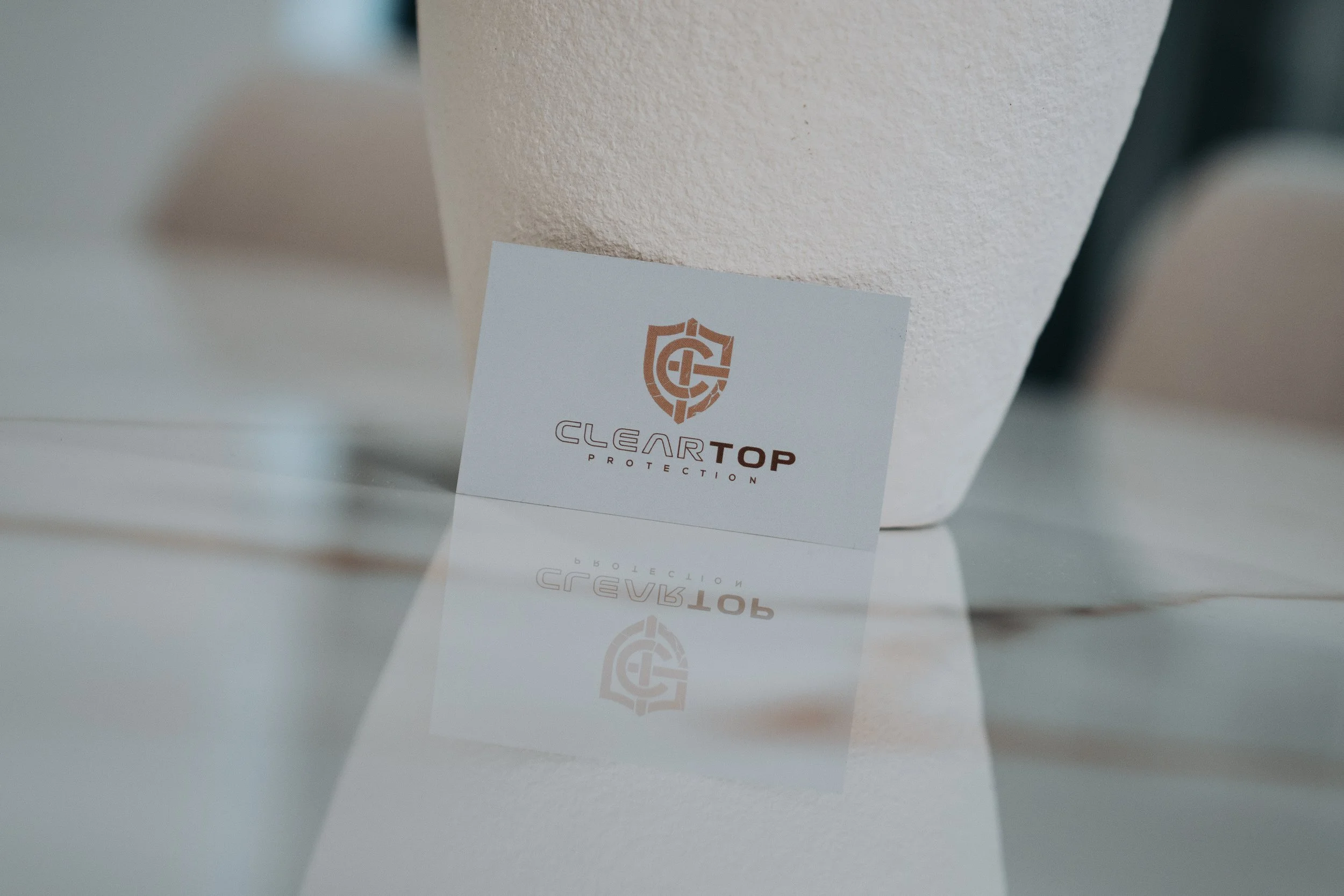 A business card with the logo and name 'Clear Top Protection' placed on a white surface with a shot of a white textured wall in the background.