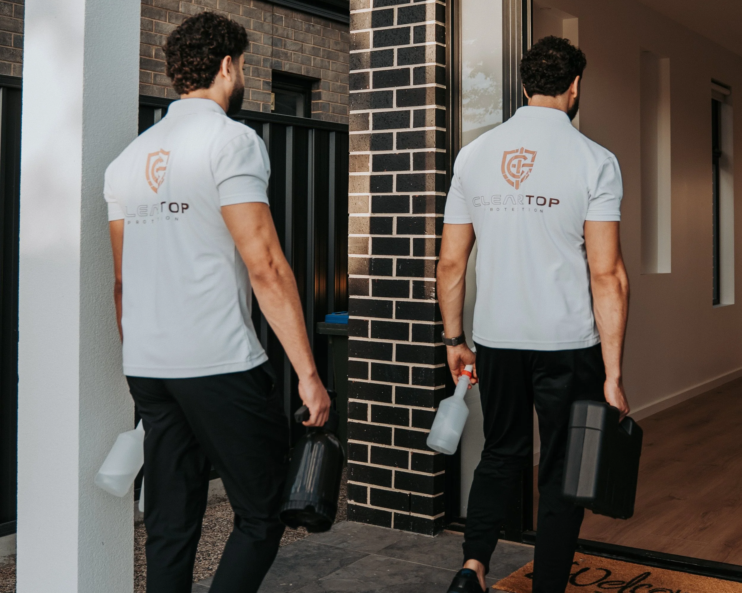 Two pest control professionals in white shirts with a shield logo on the back, carrying spray bottles and equipment, entering a house through a glass door with a brick wall and interior hallway visible.