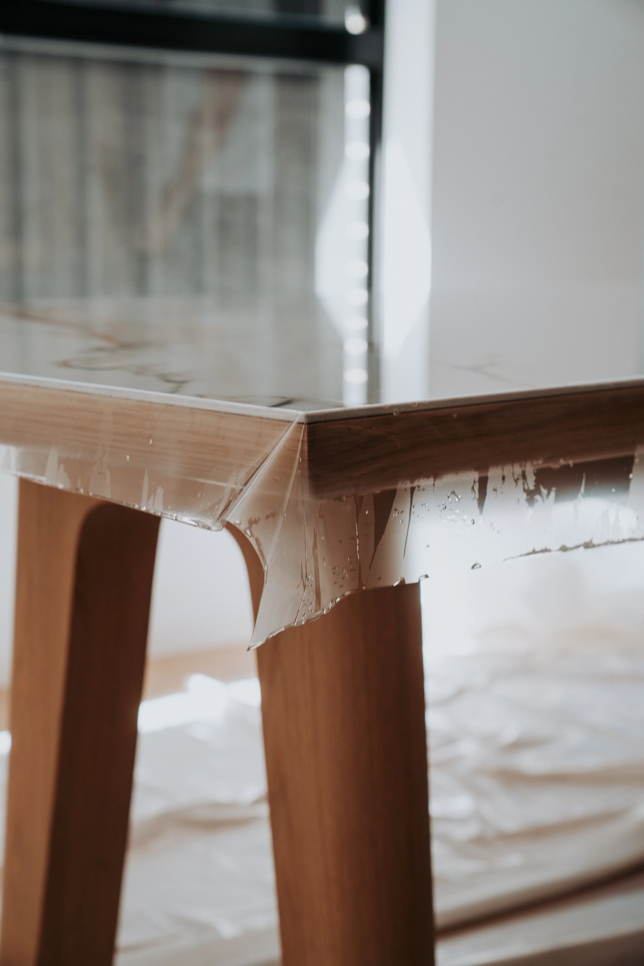 Close-up view of a wooden table with a clear plastic tablecloth that is slightly wrinkled. In the background, part of a metal appliance or equipment is visible.