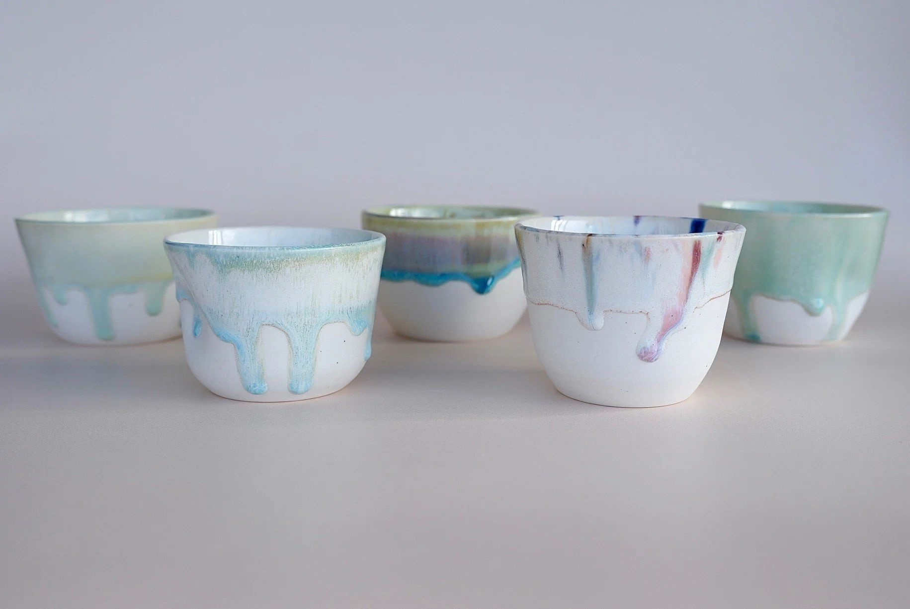 Five white ceramic bowls with pastel-colored glaze drips on the outside, arranged on a plain white surface against a plain background.
