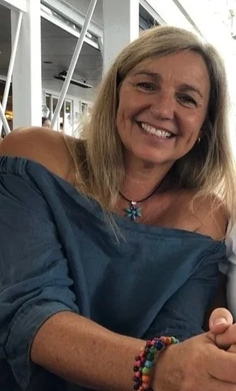 Woman smiling, wearing a blue off-shoulder top and colorful bracelets, sitting indoors.