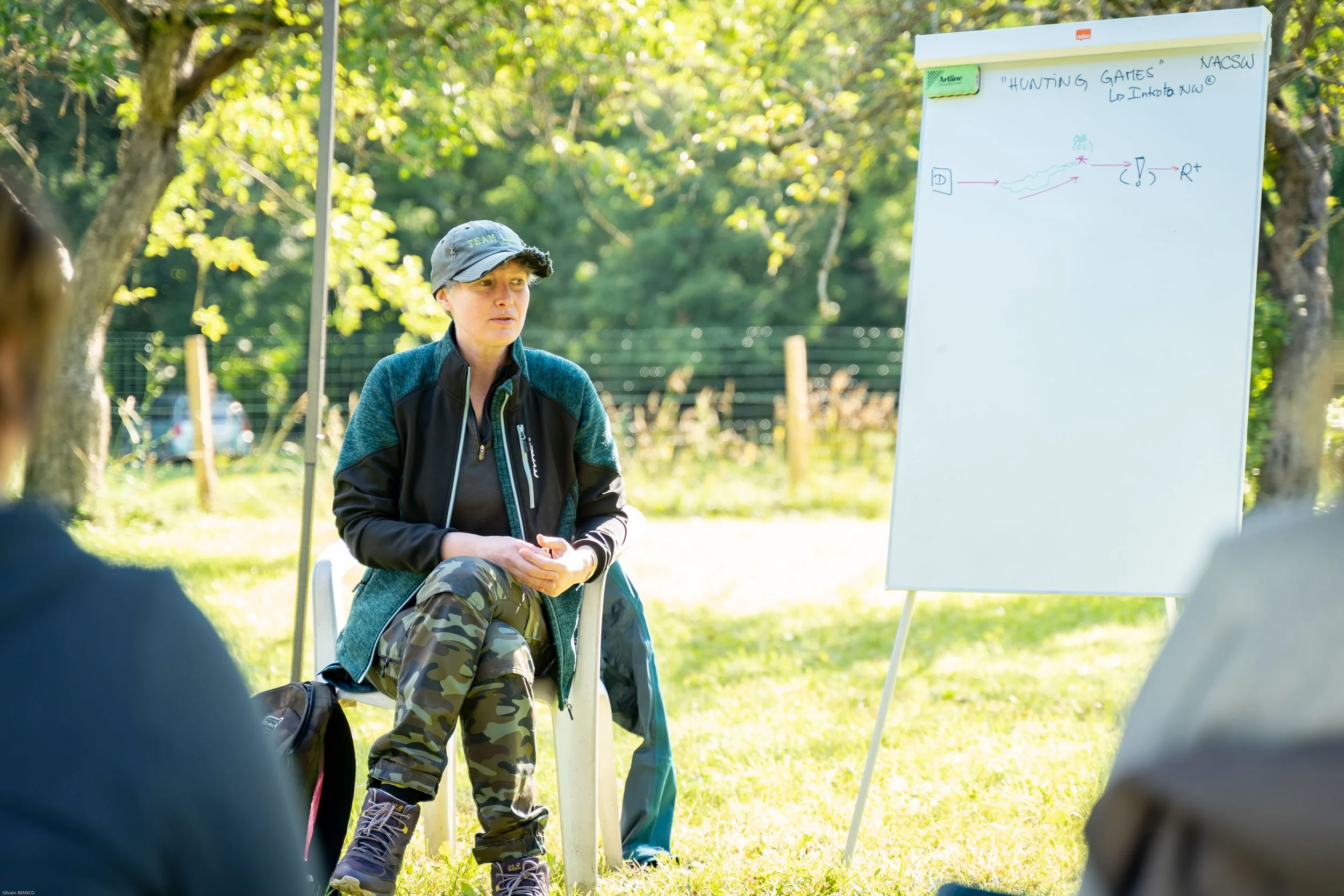 un coach en nosework : Une femme assise en plein air devant un tableau blanc avec des notes écrites, dans un environnement verdoyant, lors d'une session de formation ou de discussion.