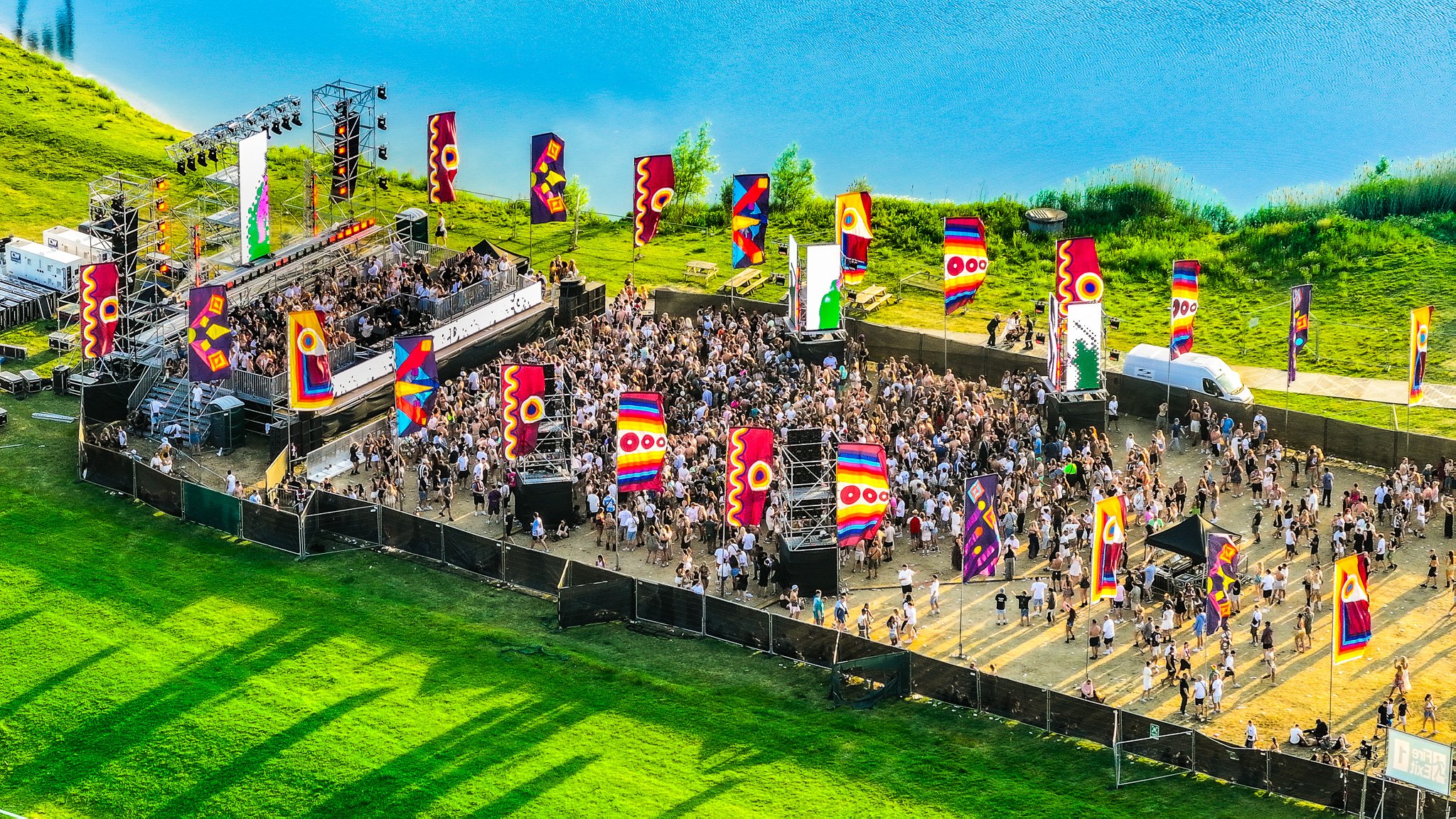 An outdoor music festival with a stage, colorful flags, and a large crowd of people, set in a grassy area near a body of water. Runway Festival 2026, Chelmsford City Race Course
