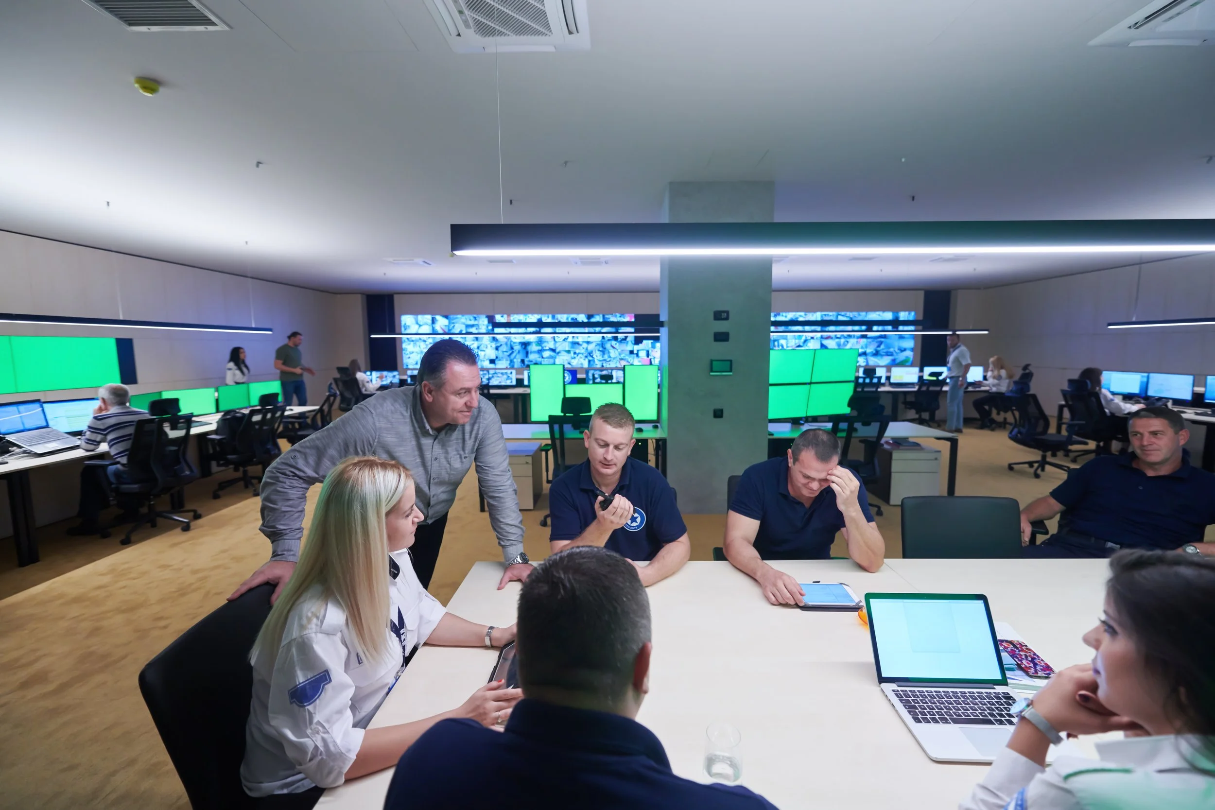 A group of people in a modern office conference room discussing and working on laptops and tablets. The room has multiple computer stations and wall screens displaying data.