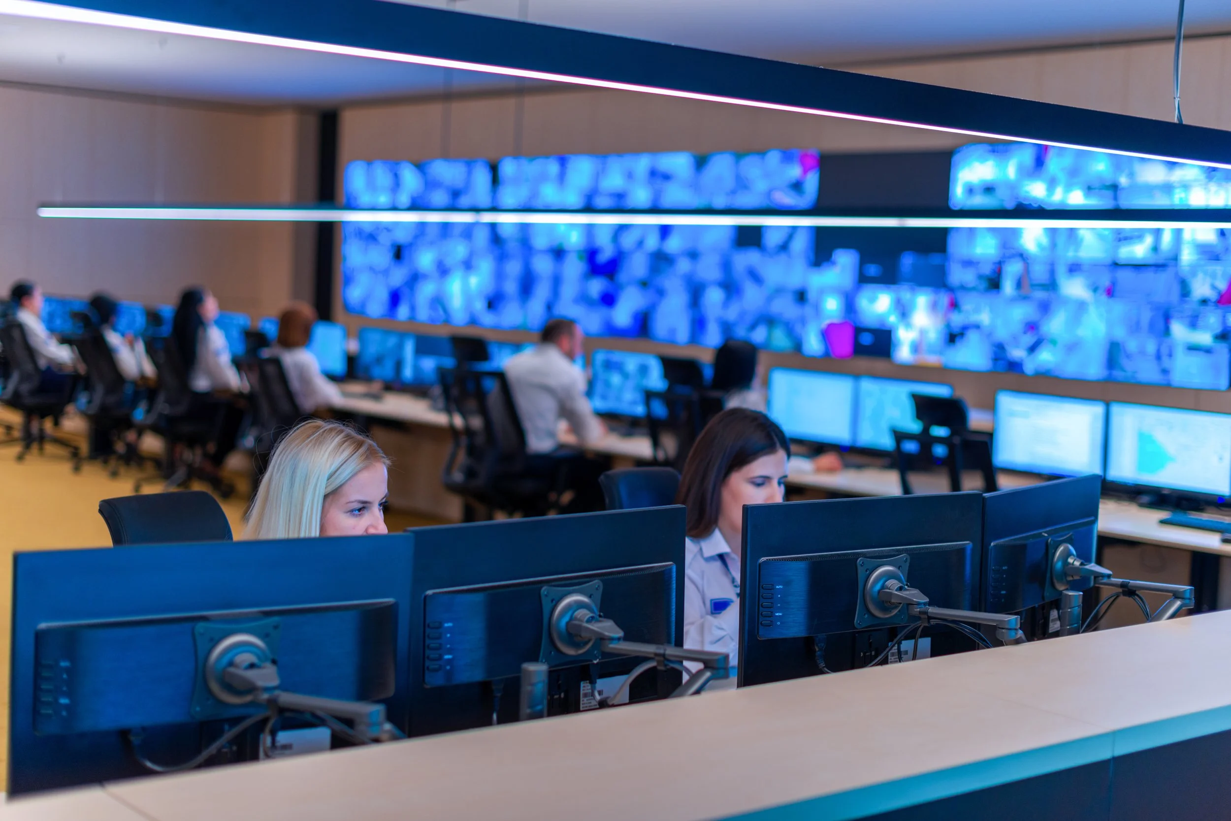 A control room with multiple people working on computers, large screens displaying data or images, illuminated with blue lighting.