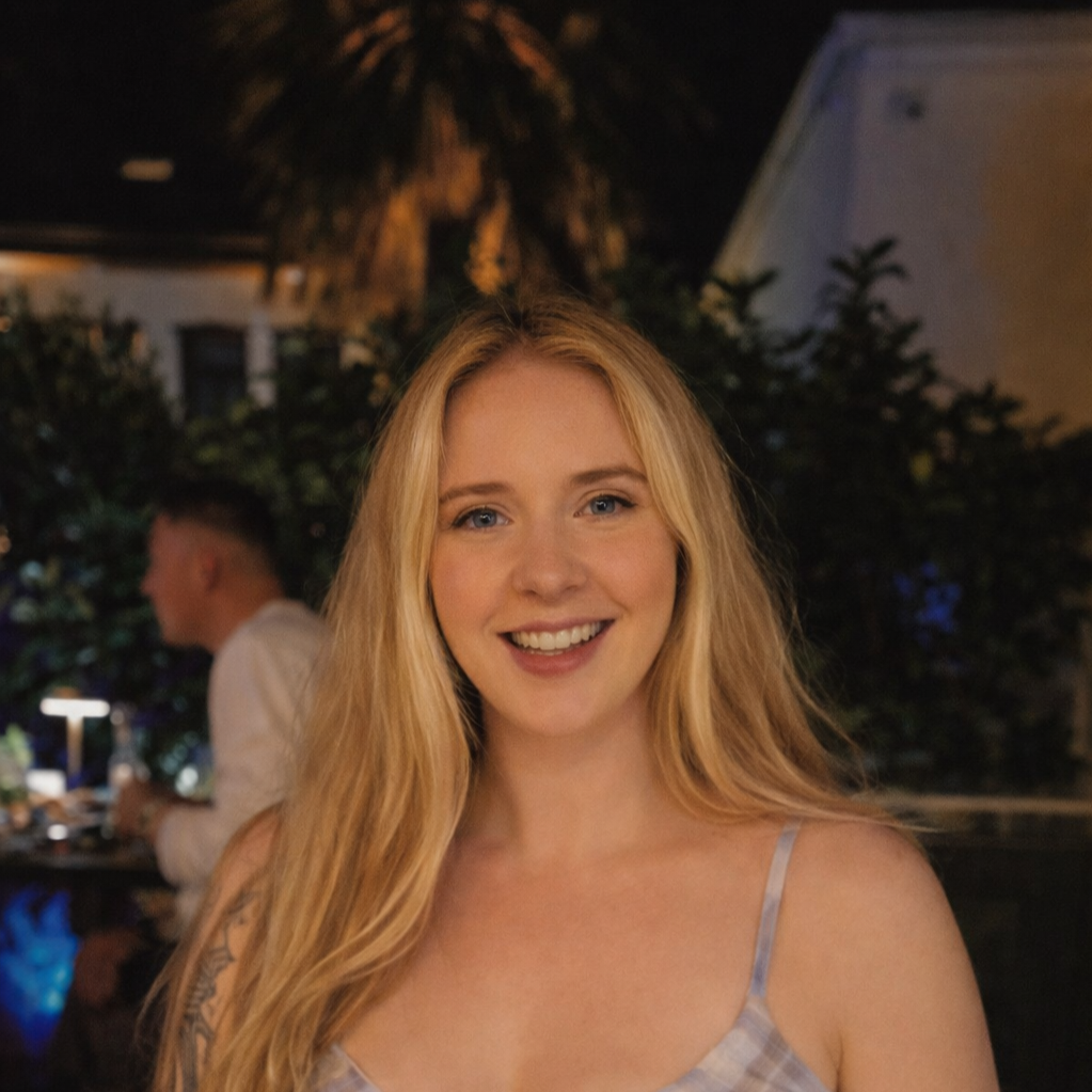 A young woman with long blonde hair smiling at the camera at night outdoor setting, with blurred background of a person, trees, and buildings.