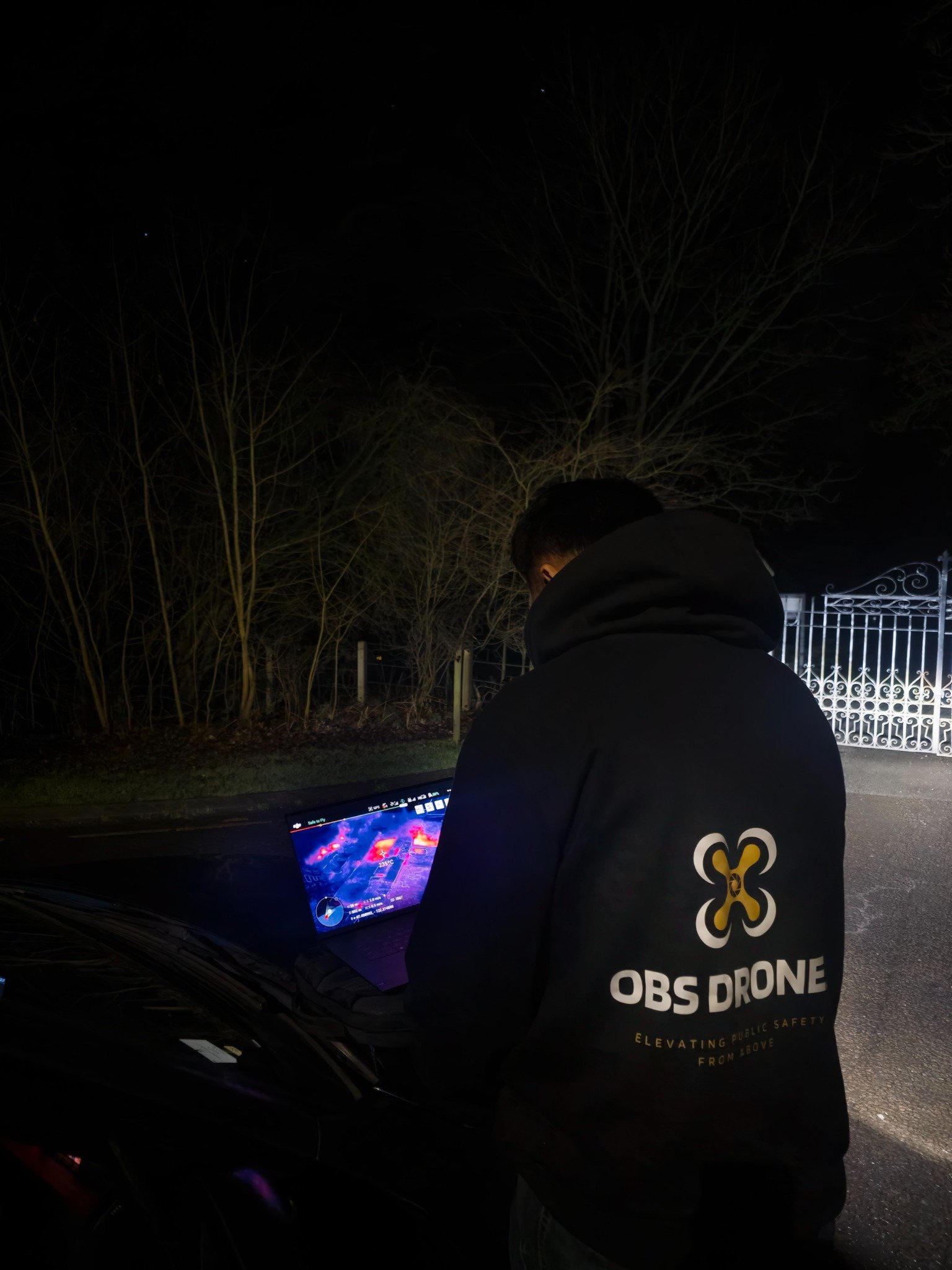 Person wearing a black jacket with yellow and white text on the back, standing outside at night, working on a drone controller with a screen displaying a thermal imaging view.