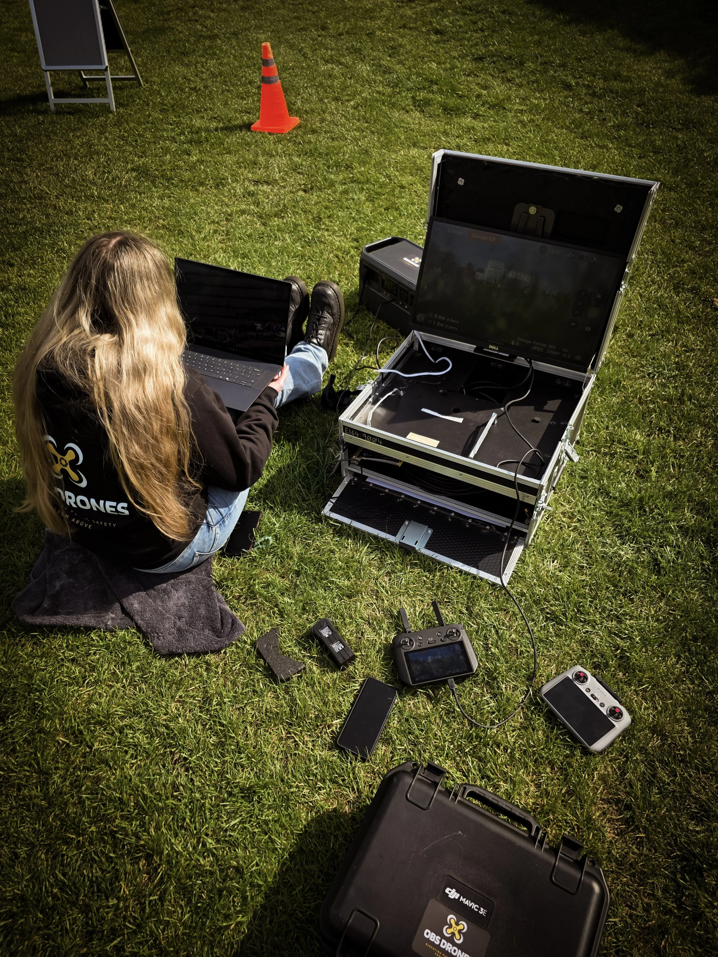 A person with long blonde hair seated on grass using a laptop, surrounded by drone equipment, including a black case with a drone logo, remote controllers, and a large screen. A traffic cone and a blackboard are also visible in the background.