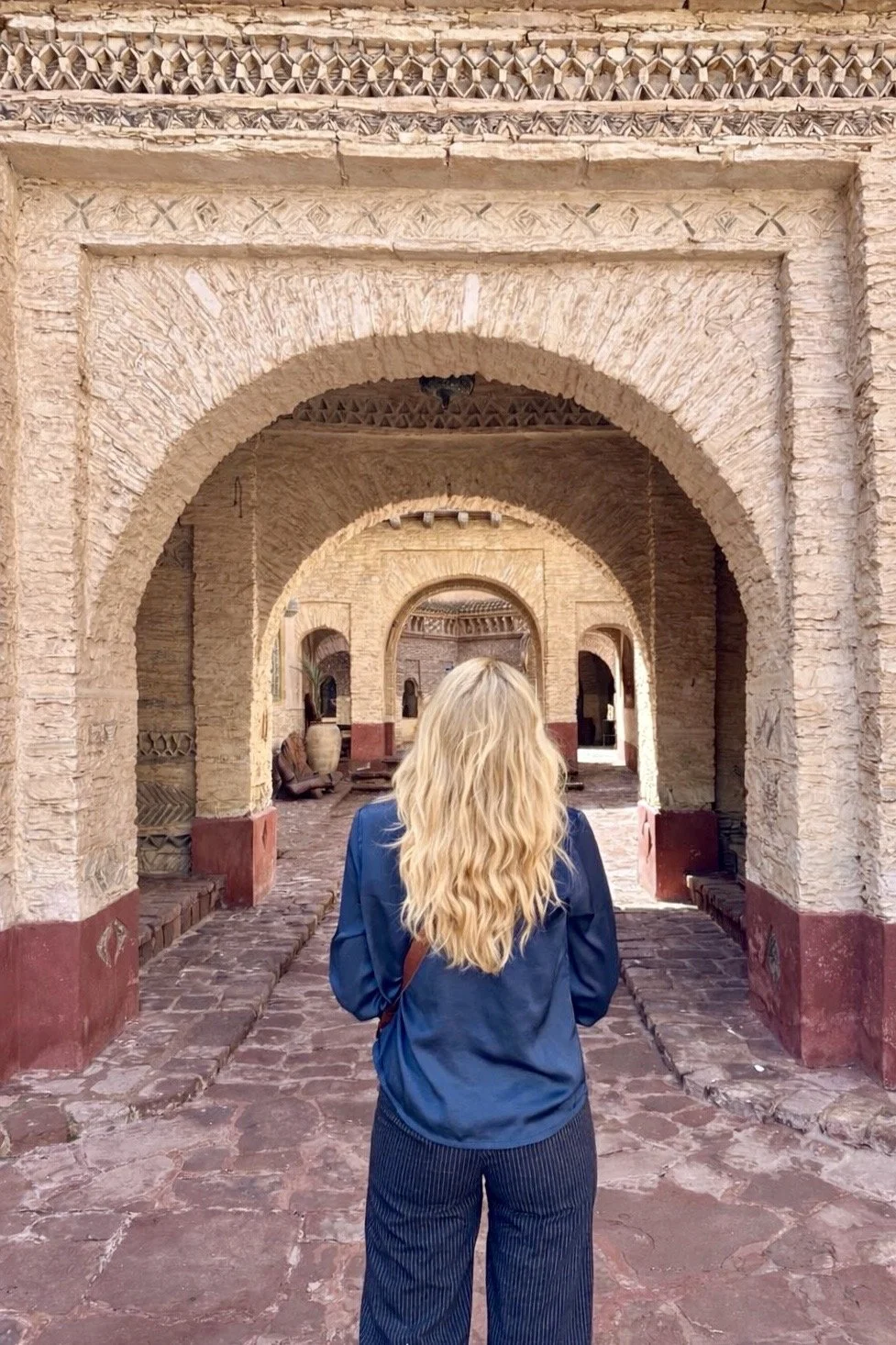 Femme aux cheveux longs et bouclés, portant une veste bleue, marche dans une cour en pierre entourée d'arches en adobe, architecture traditionnelle du Maroc.
