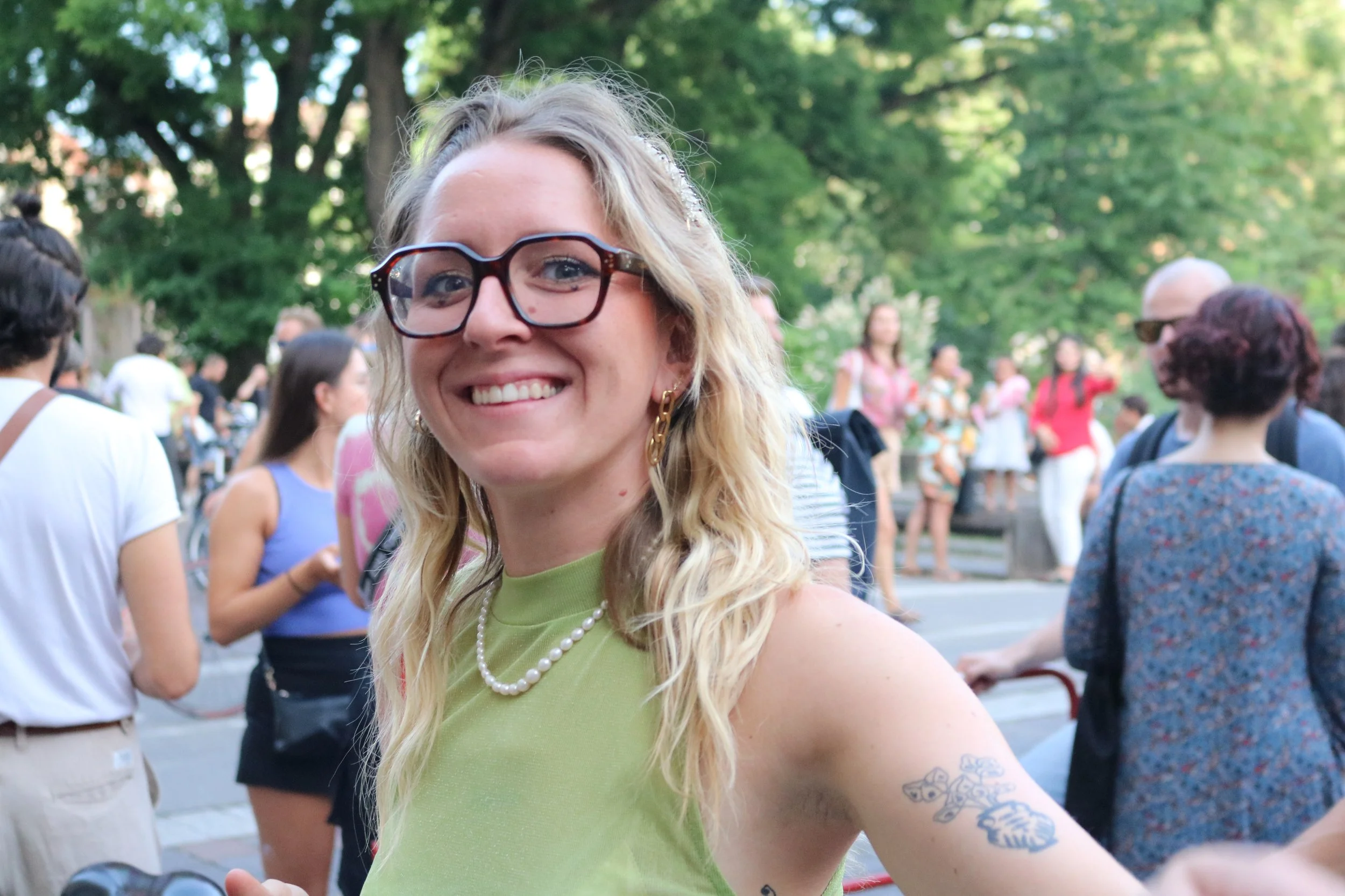 Une femme souriante portant des lunettes, une robe verte, une perle et des boucles d'oreilles, avec un tatouage à l'avant-bras, entourée d'un groupe de personnes dans un parc arboré.