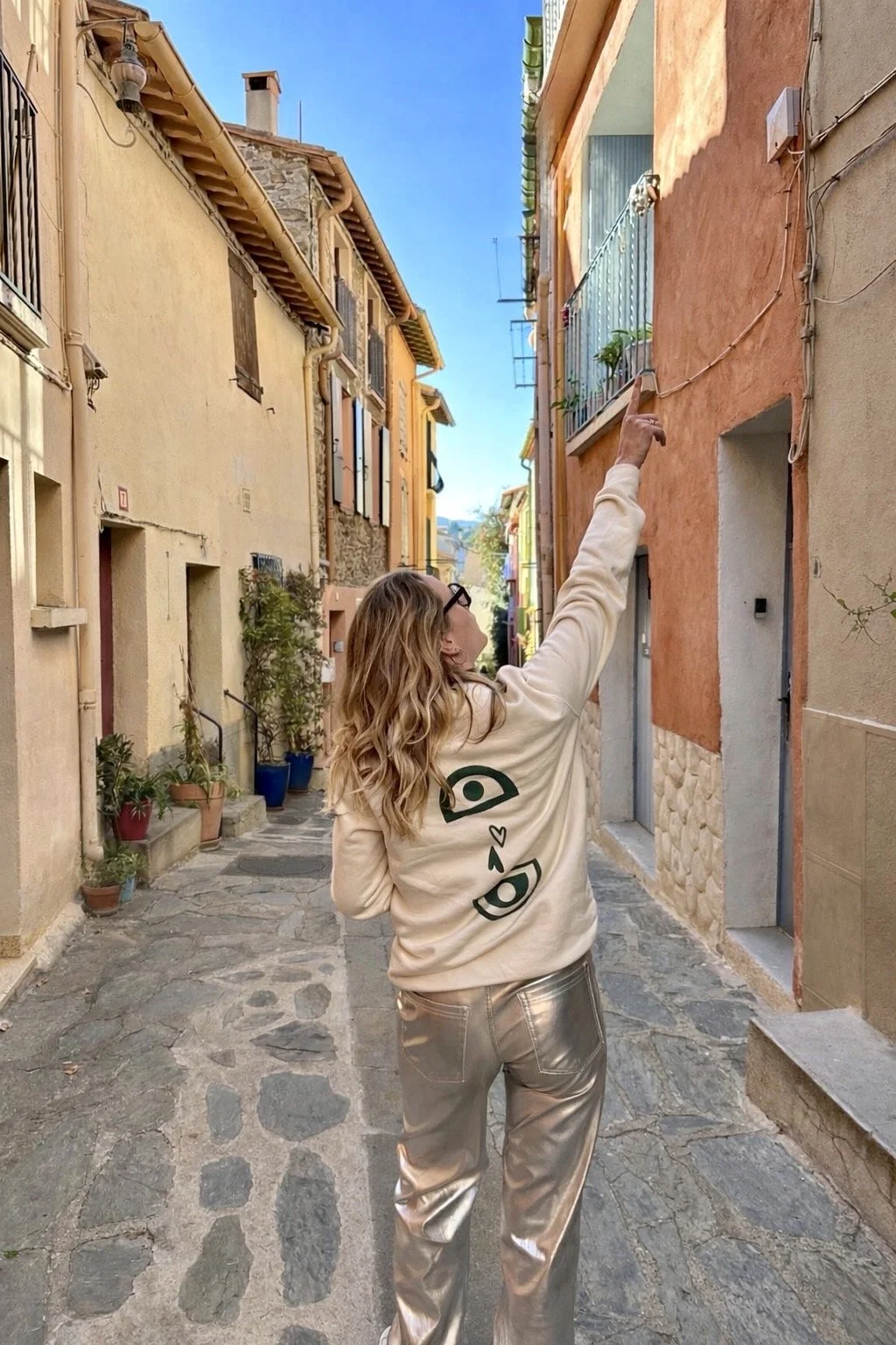 Femme avec des cheveux bouclés, portant un sweatshirt beige avec un dessin de visage et un cœur, et un pantalon en faux cuir argenté, pointe une fenêtre dans une ruelle colorée.