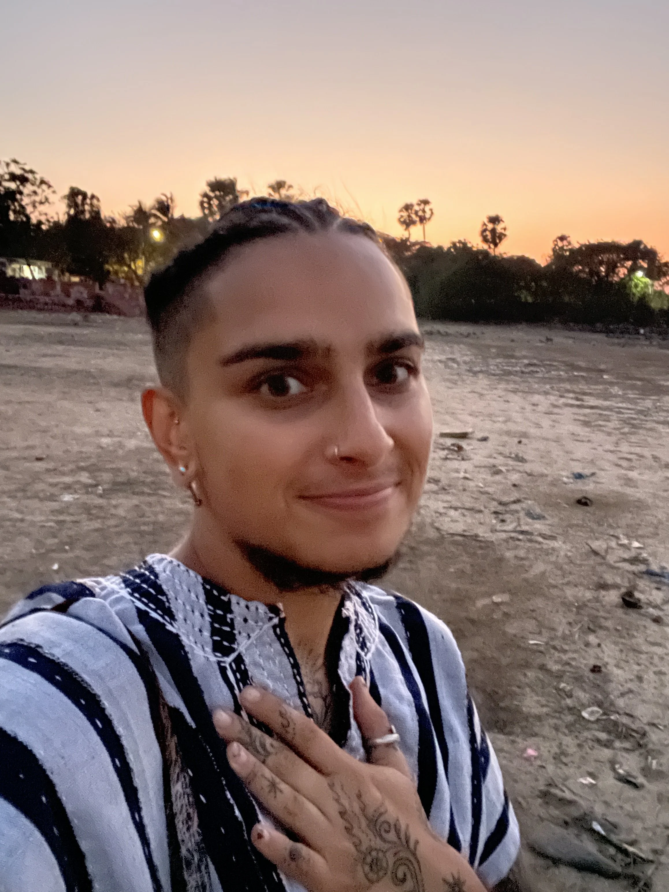 A person with dark, short hair taking a selfie on a beach during sunset, wearing blue and white striped traditional clothing  and showing a henna tattoo on their left hand.