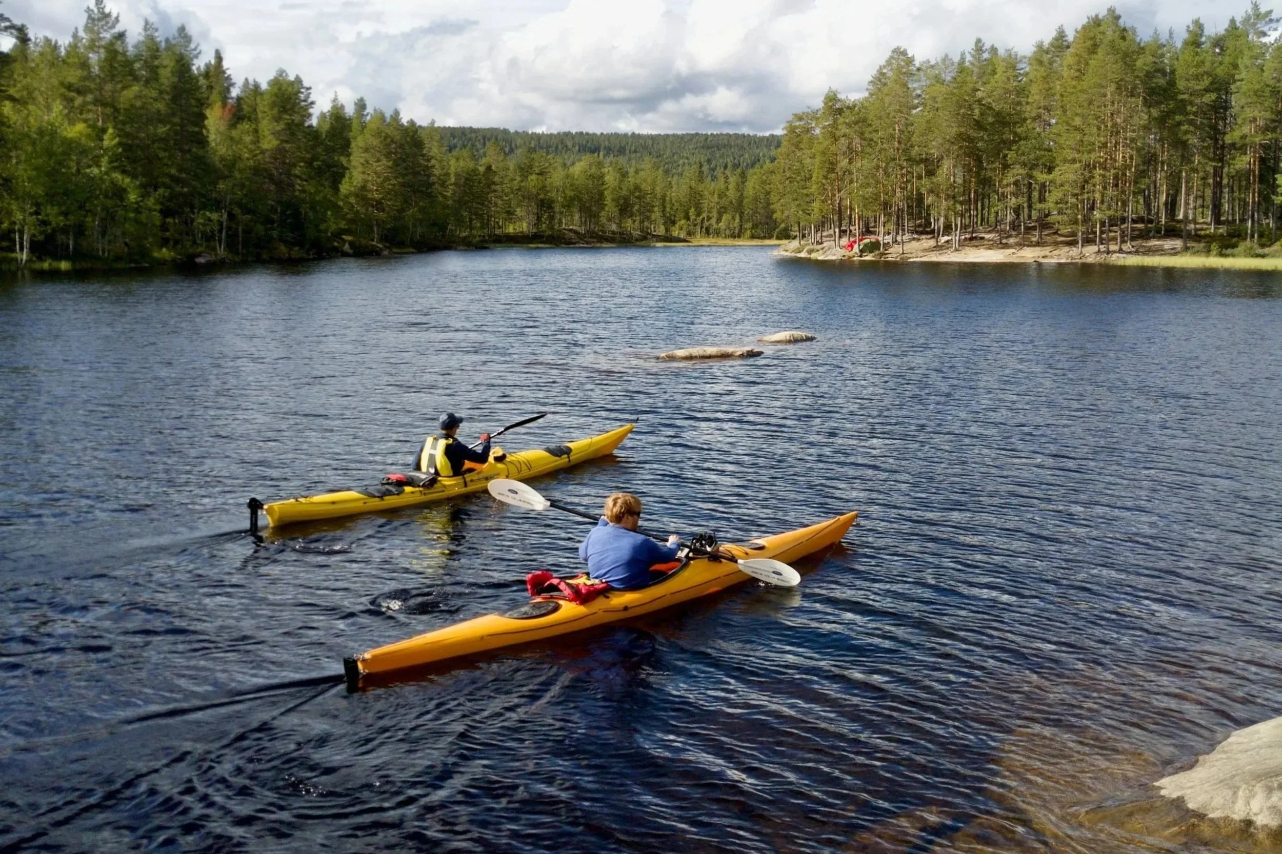 Three people kayaking on a calm lake surrounded by pine trees with a forested hillside in the background.