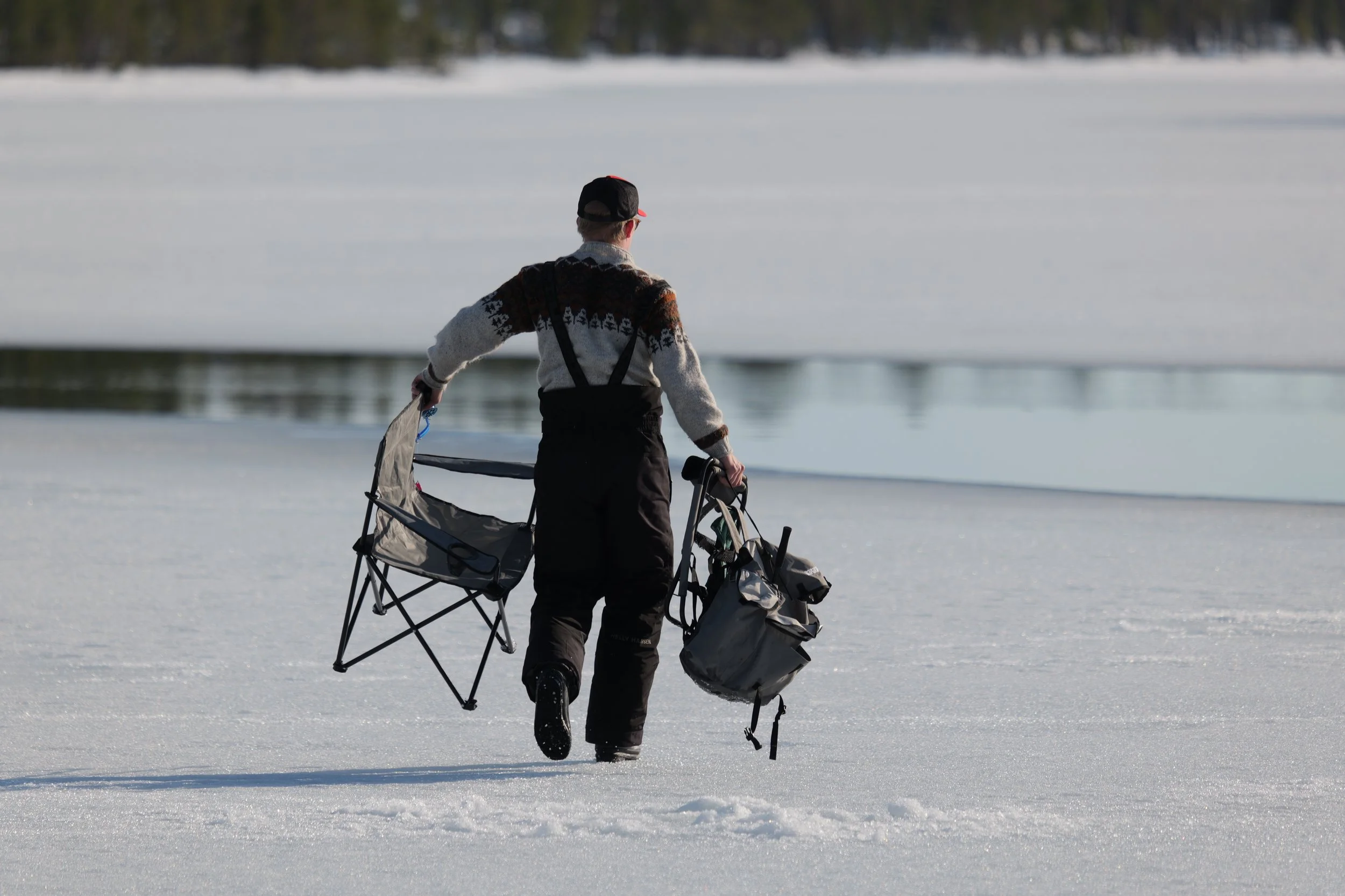 A person walking on snow-covered ground near a body of water, carrying outdoor gear and walking away from the camera on a clear day.