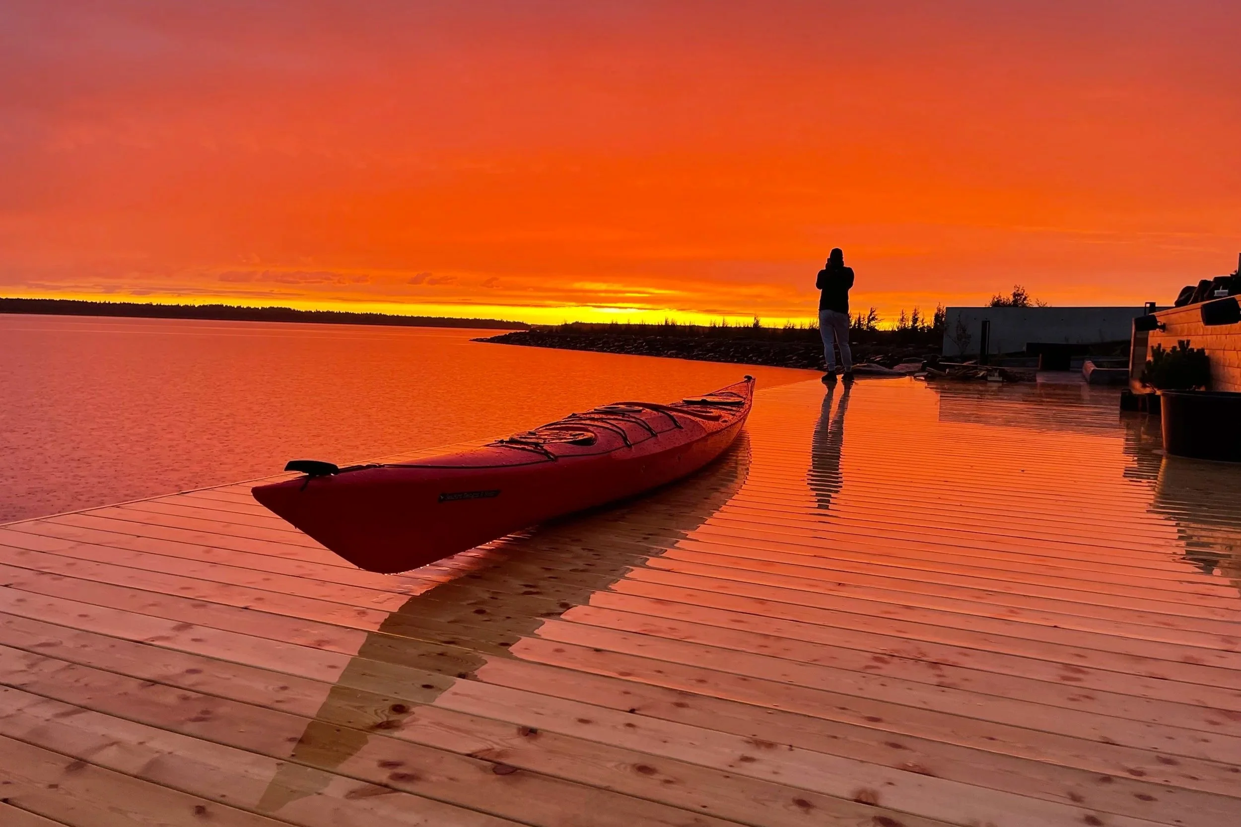 A person standing on a wooden dock at sunset, with a kayak on the dock and a body of water in the background, under an orange sky.