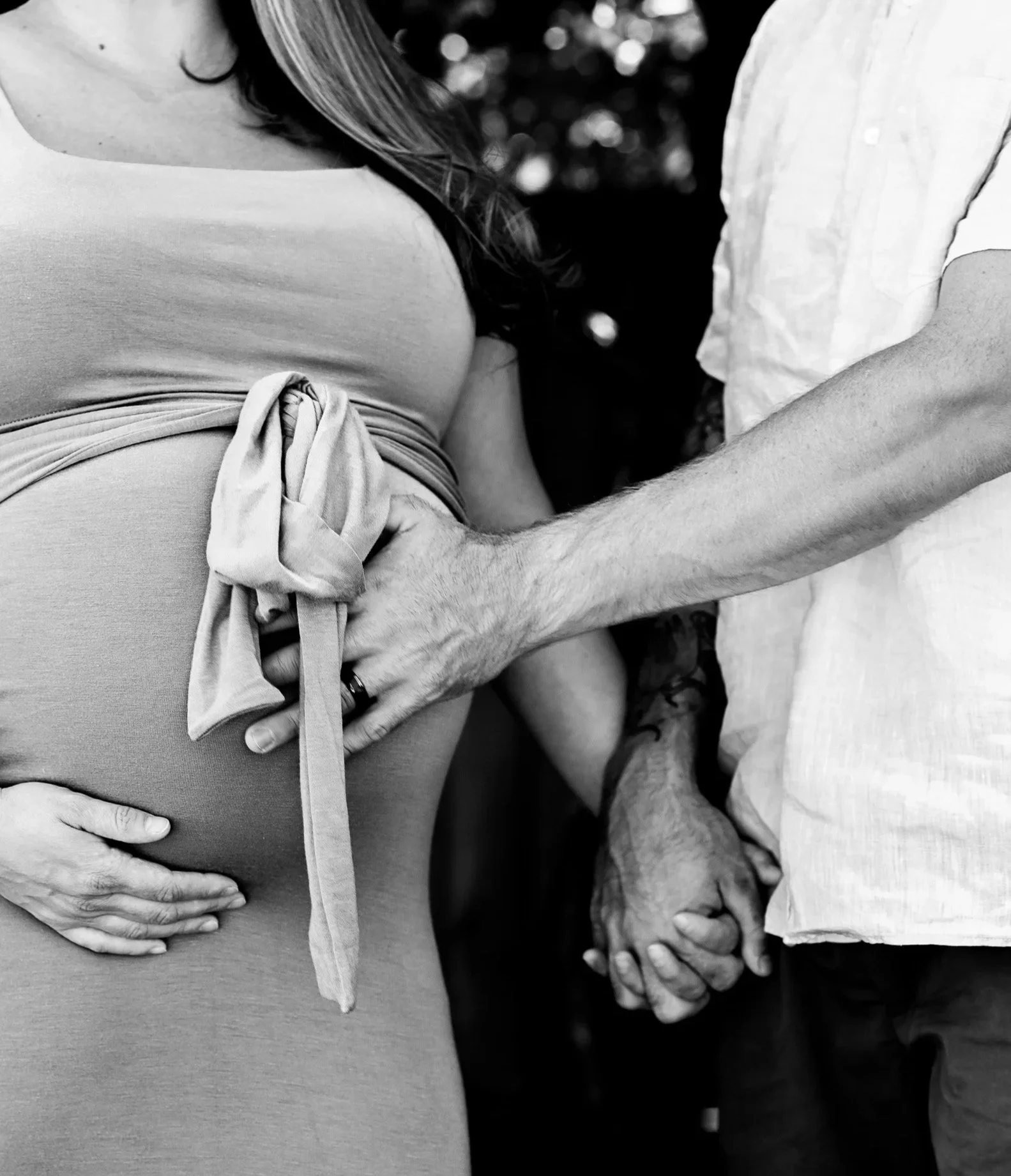Close-up black and white image of a pregnant woman with a man, both with their hands on her belly, while holding hands.