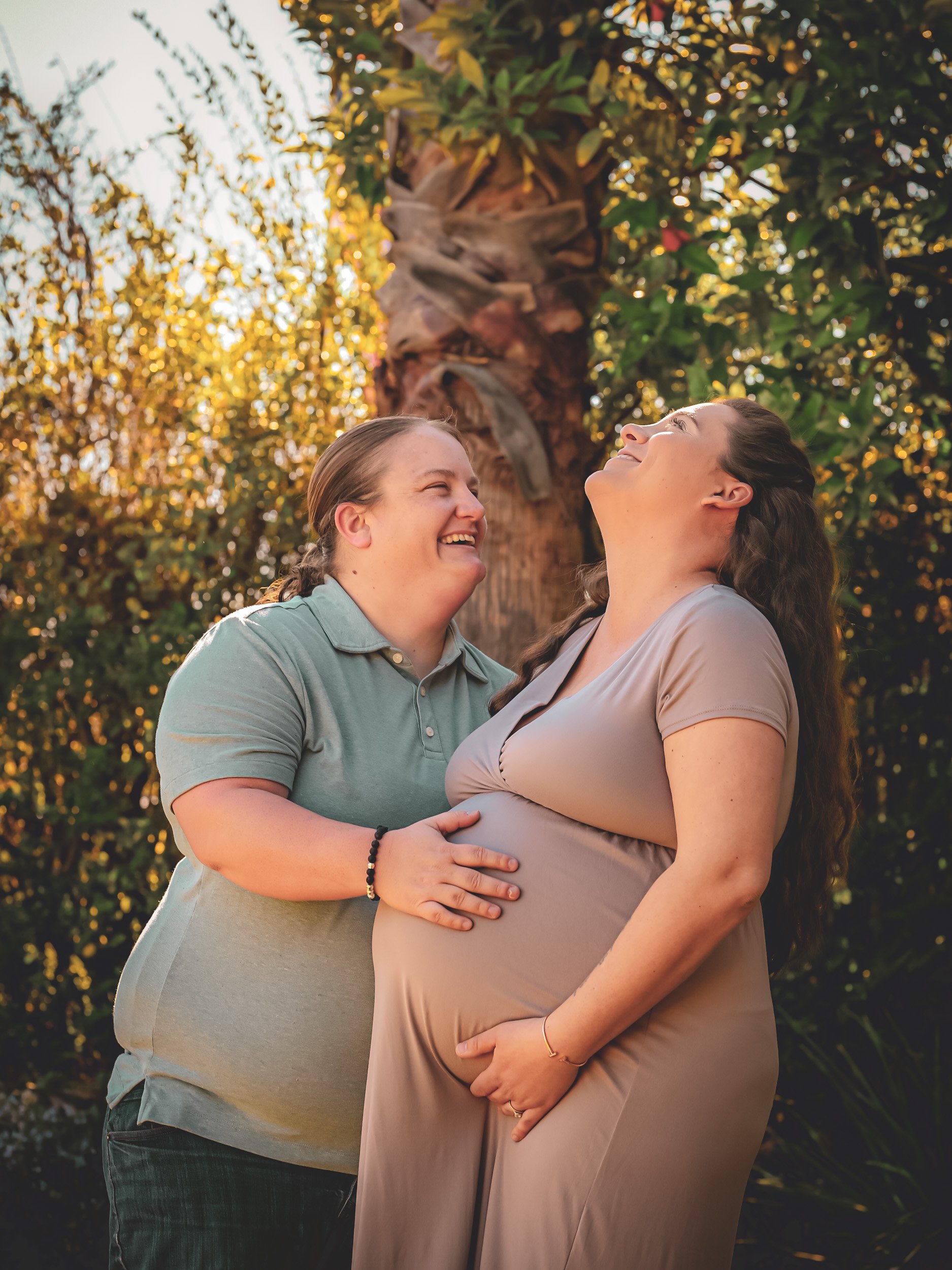 Two women smiling and looking at each other outdoors during sunset, with a pregnant woman holding her belly, in front of a lush green tree.